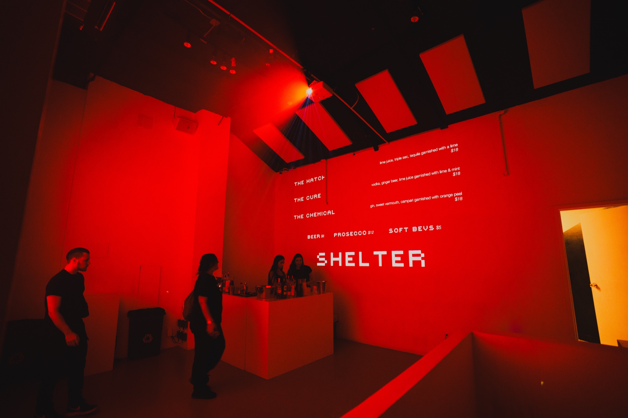 A red lit room with a bar and a cocktail menu projected above the word Shelter in white letters