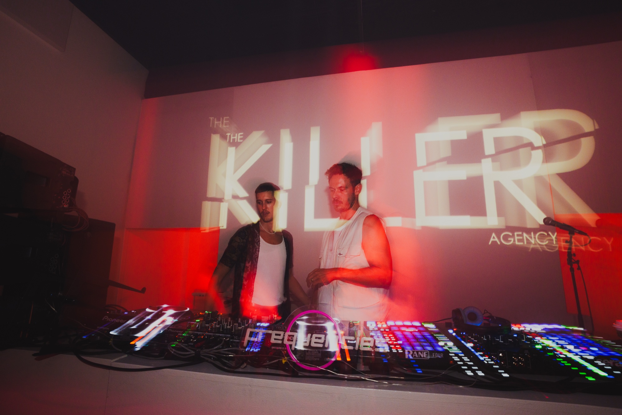 Two DJs at a glowing console beneath a red The Killer Agency wall projection in New York City