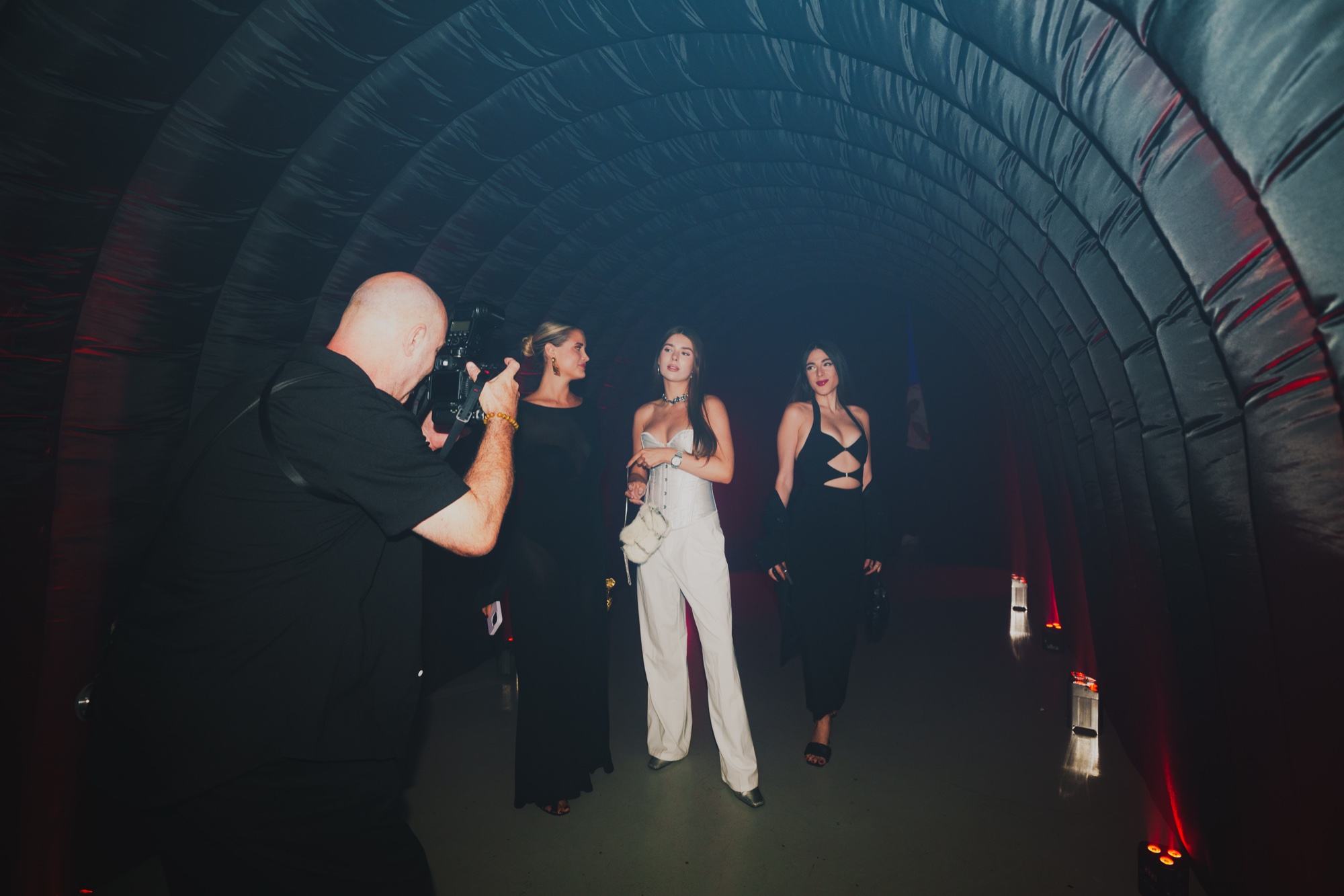 Photographer shooting three women in evening wear walking through a dark quilted tunnel