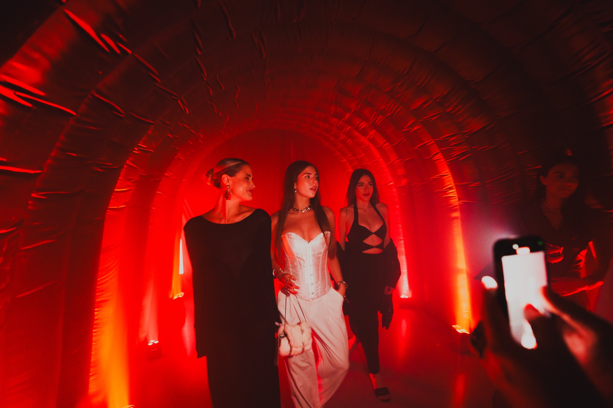 Three women walk through a red lit fabric tunnel, one in a white corset dress, a phone raised in front