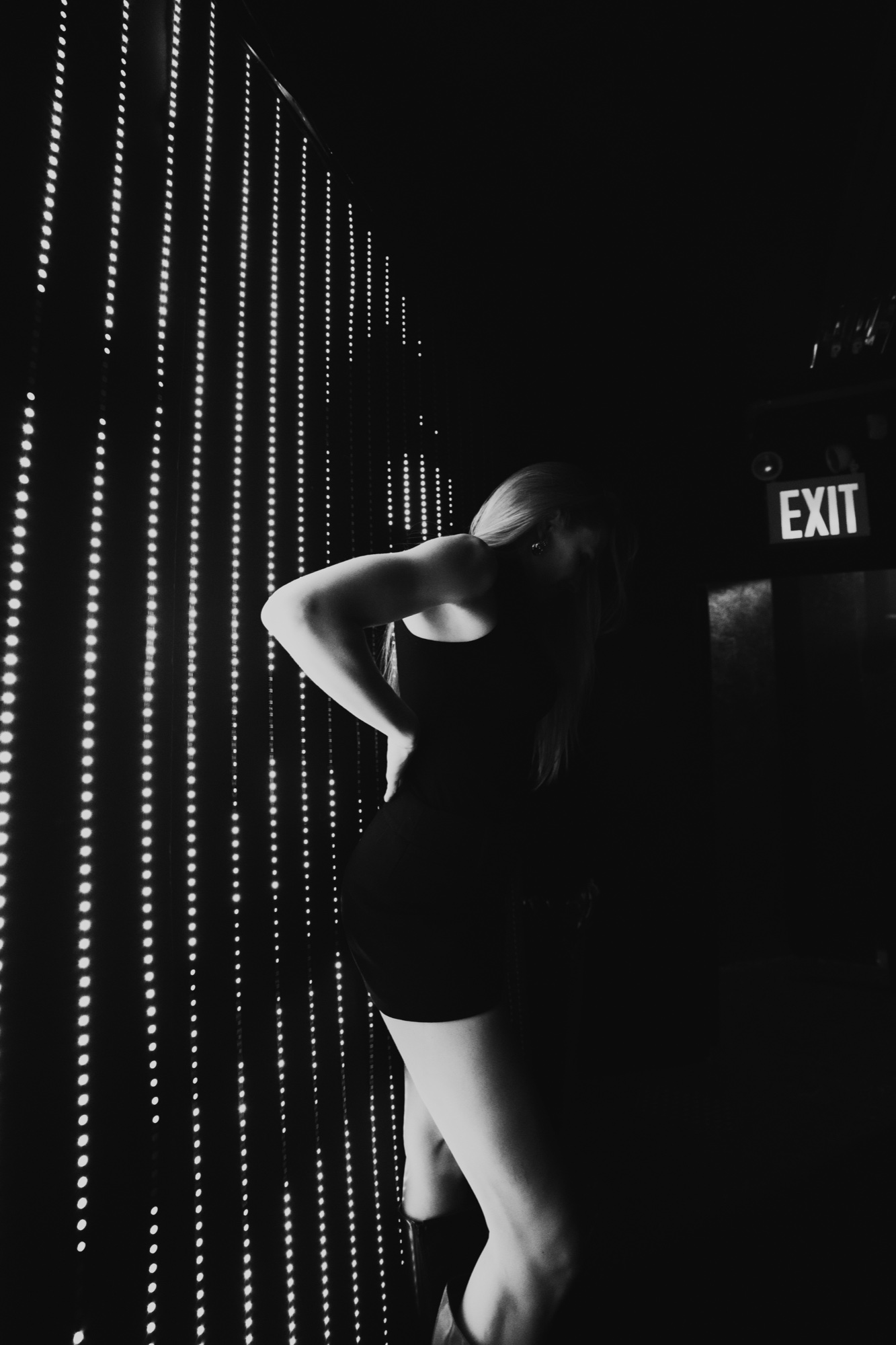 Monochrome silhouette of a woman in a fitted dress beside a wall of dotted light and an exit sign