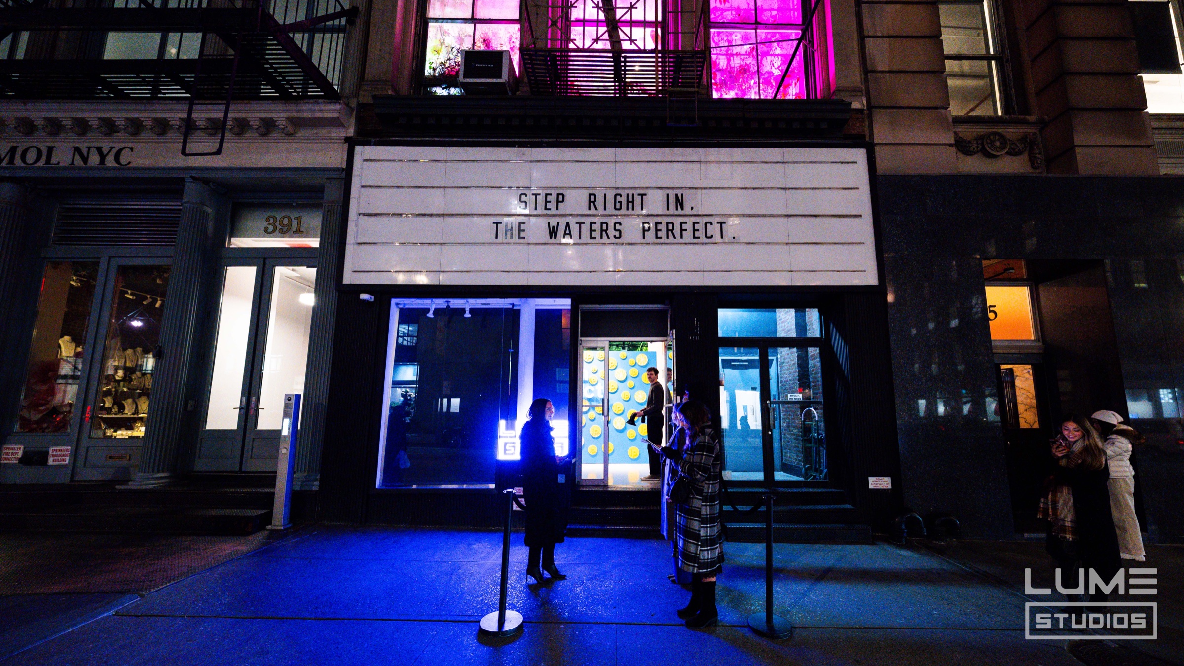 Marquee reads “step right in, the water’s perfect” above a glowing blue sidewalk at night
