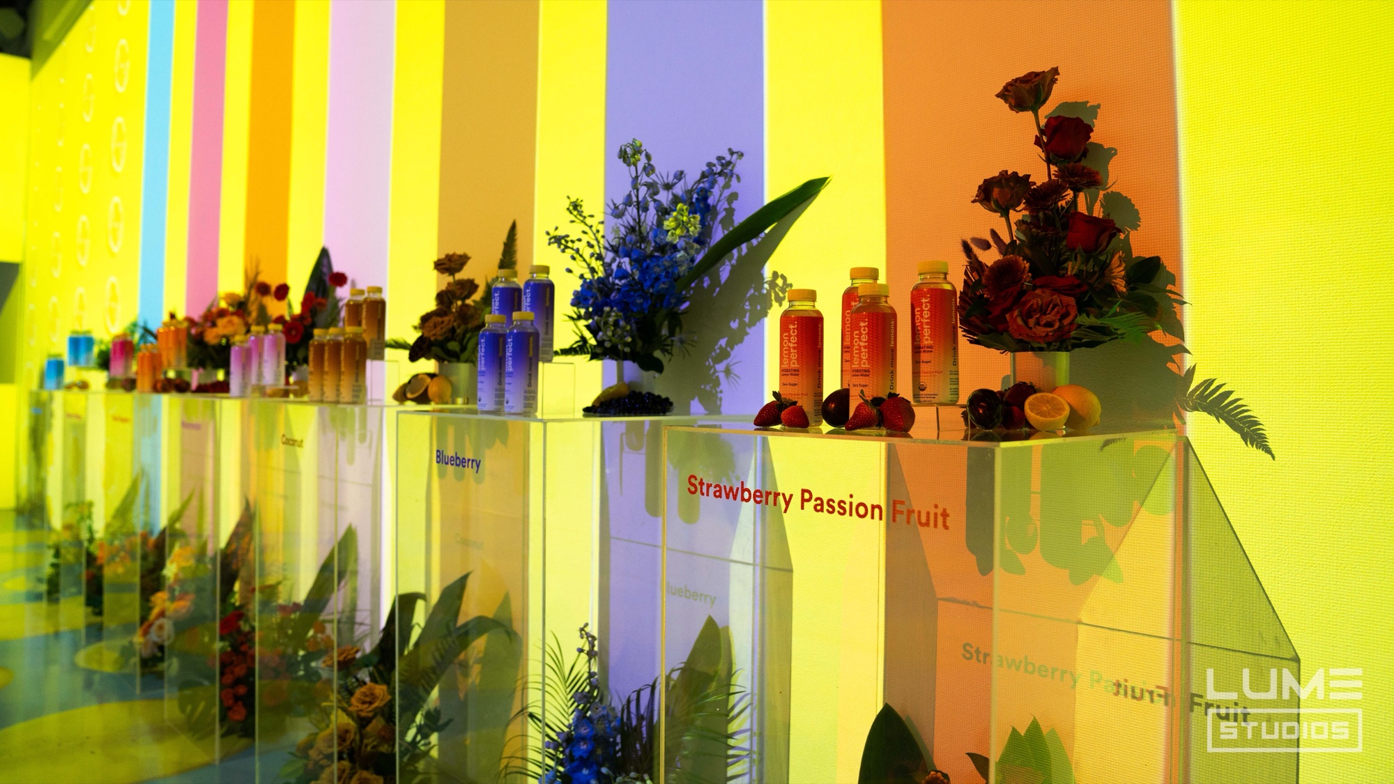 Acrylic flavor displays with Lemon Perfect bottles, fruit, and flowers against striped projections
