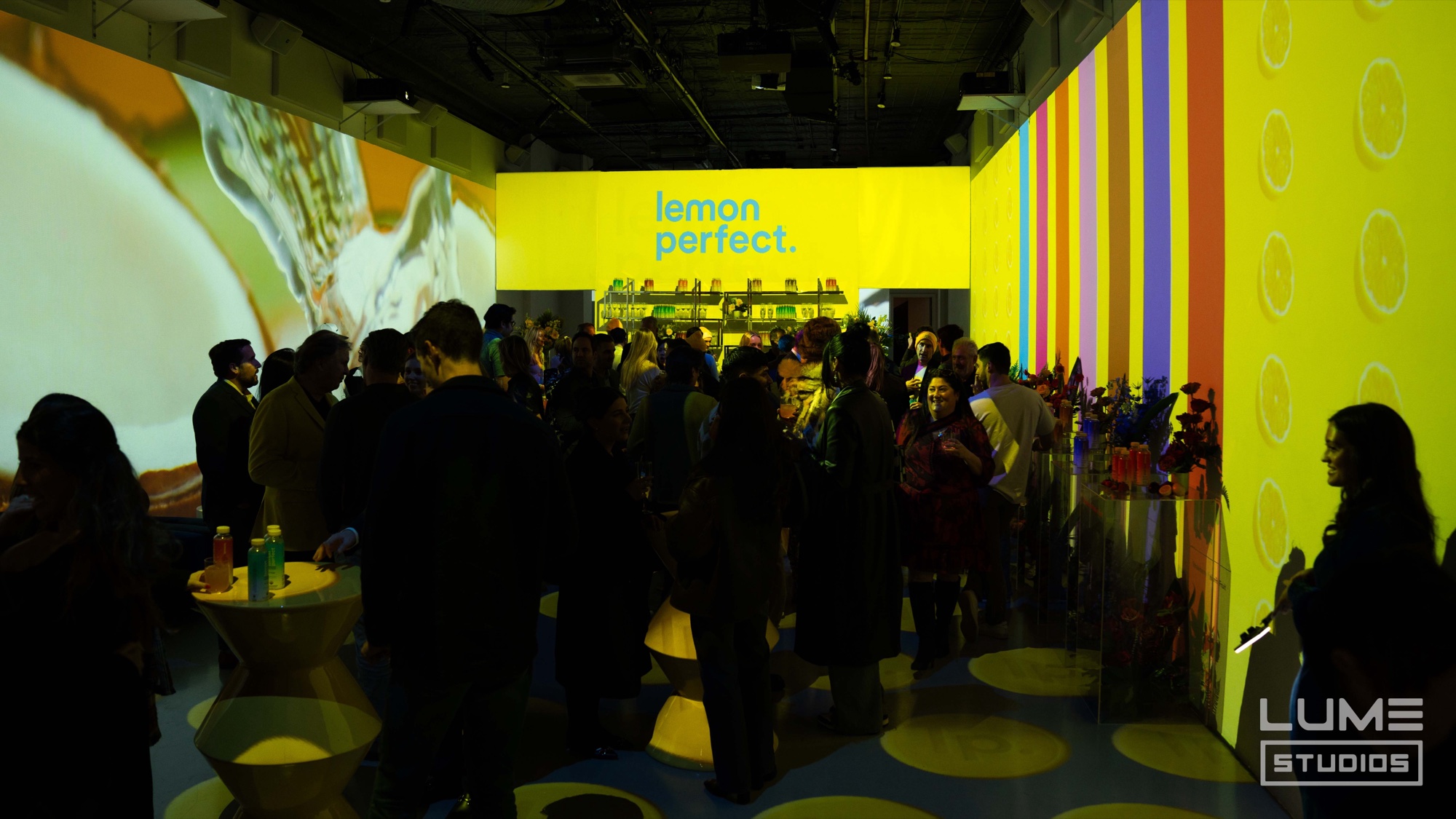 Crowd mingles under a glowing Lemon Perfect logo with striped and citrus projection walls
