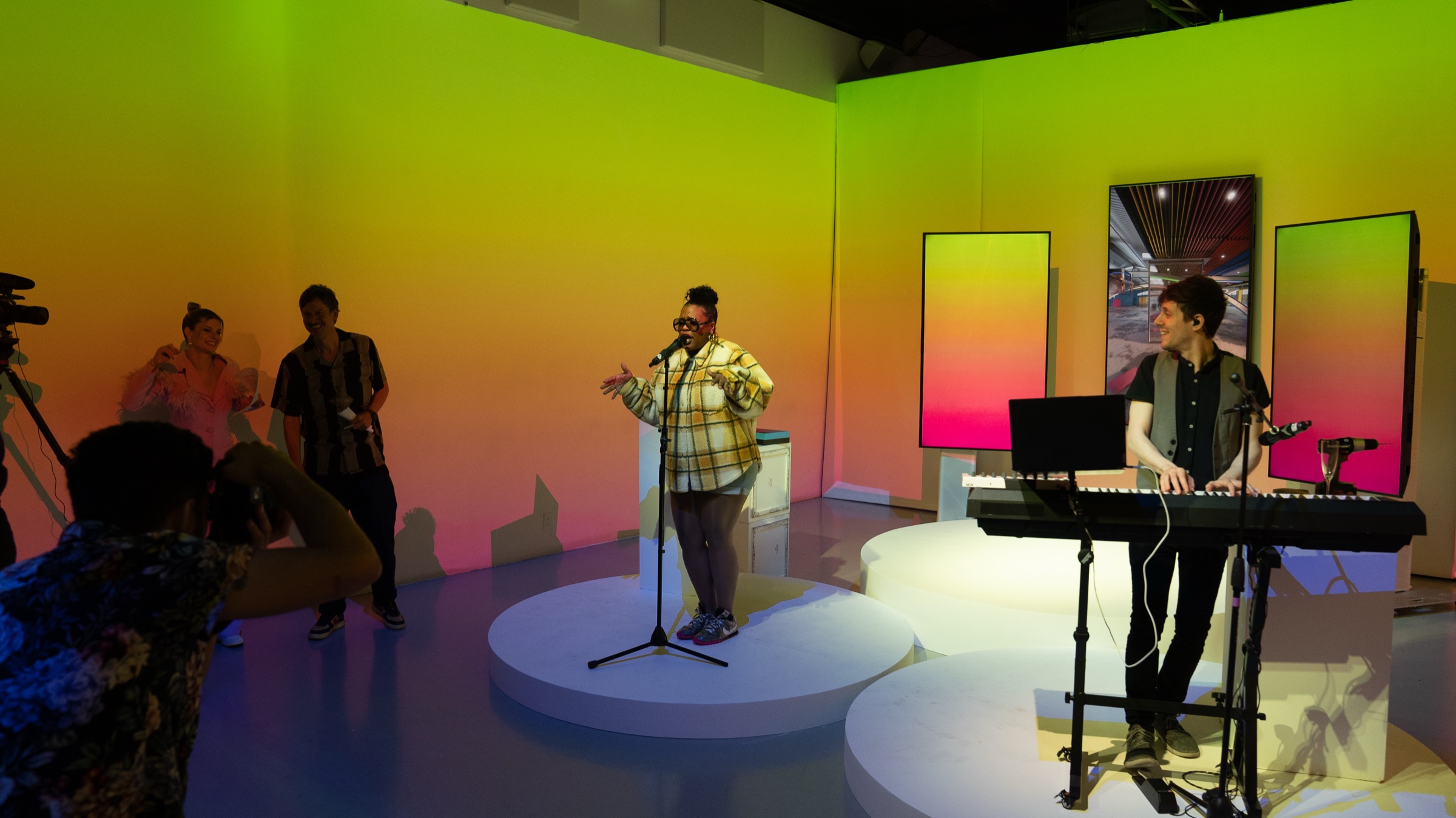 A singer in a plaid shirt and a keyboardist perform on a yellow and pink gradient set while filmed
