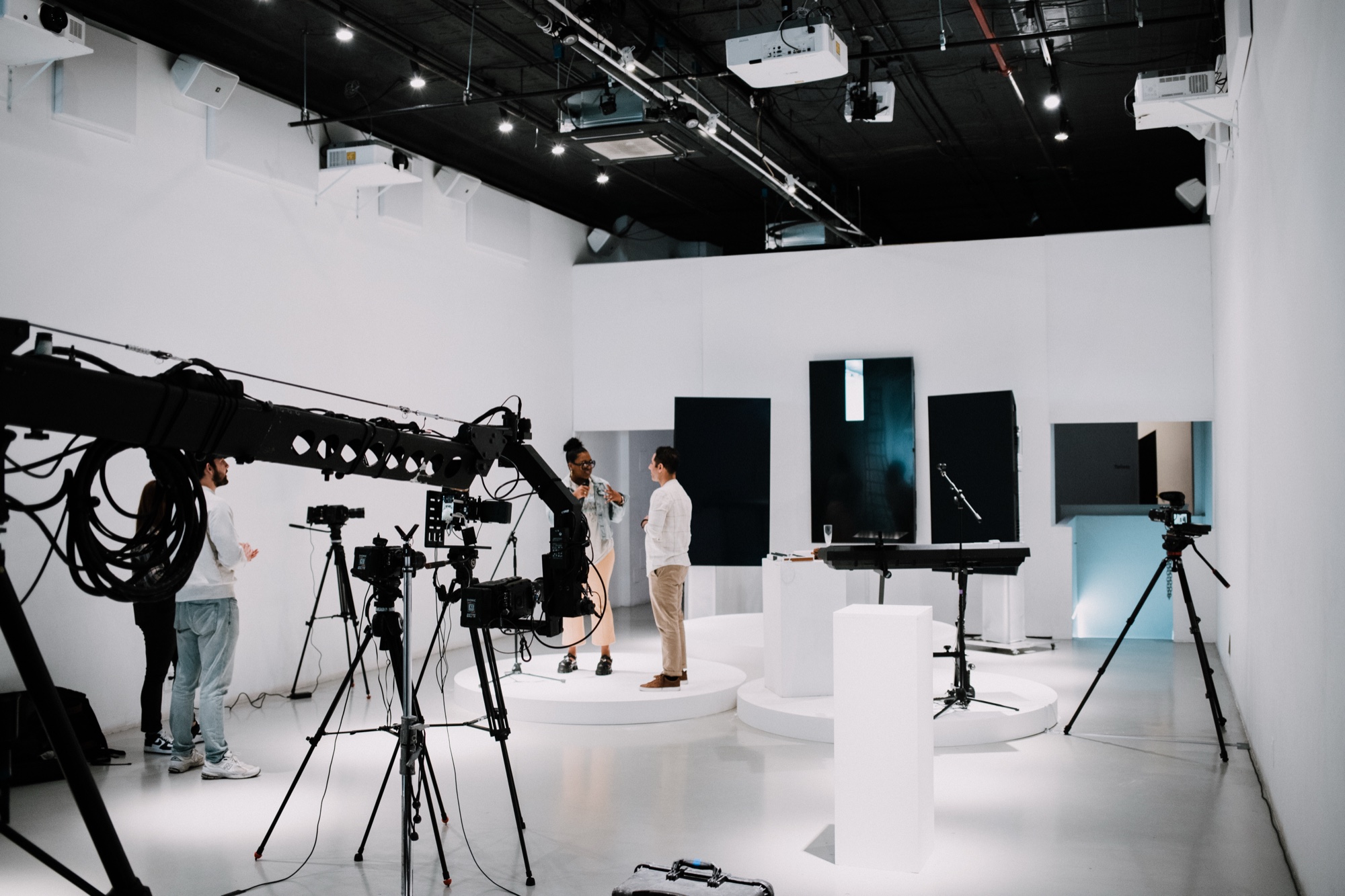 Camera crane and tripods aimed at two presenters on a round white stage in a white studio