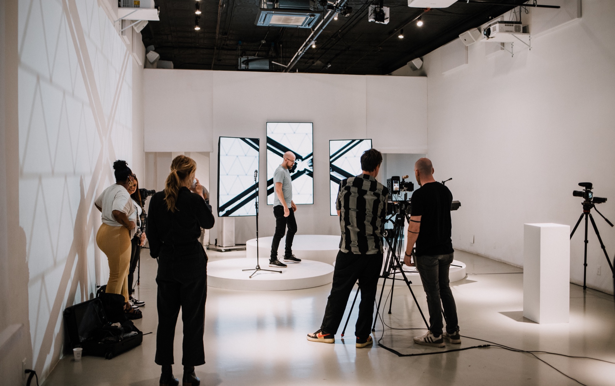 A crew films a speaker standing on a round white platform between vertical screens in a white studio