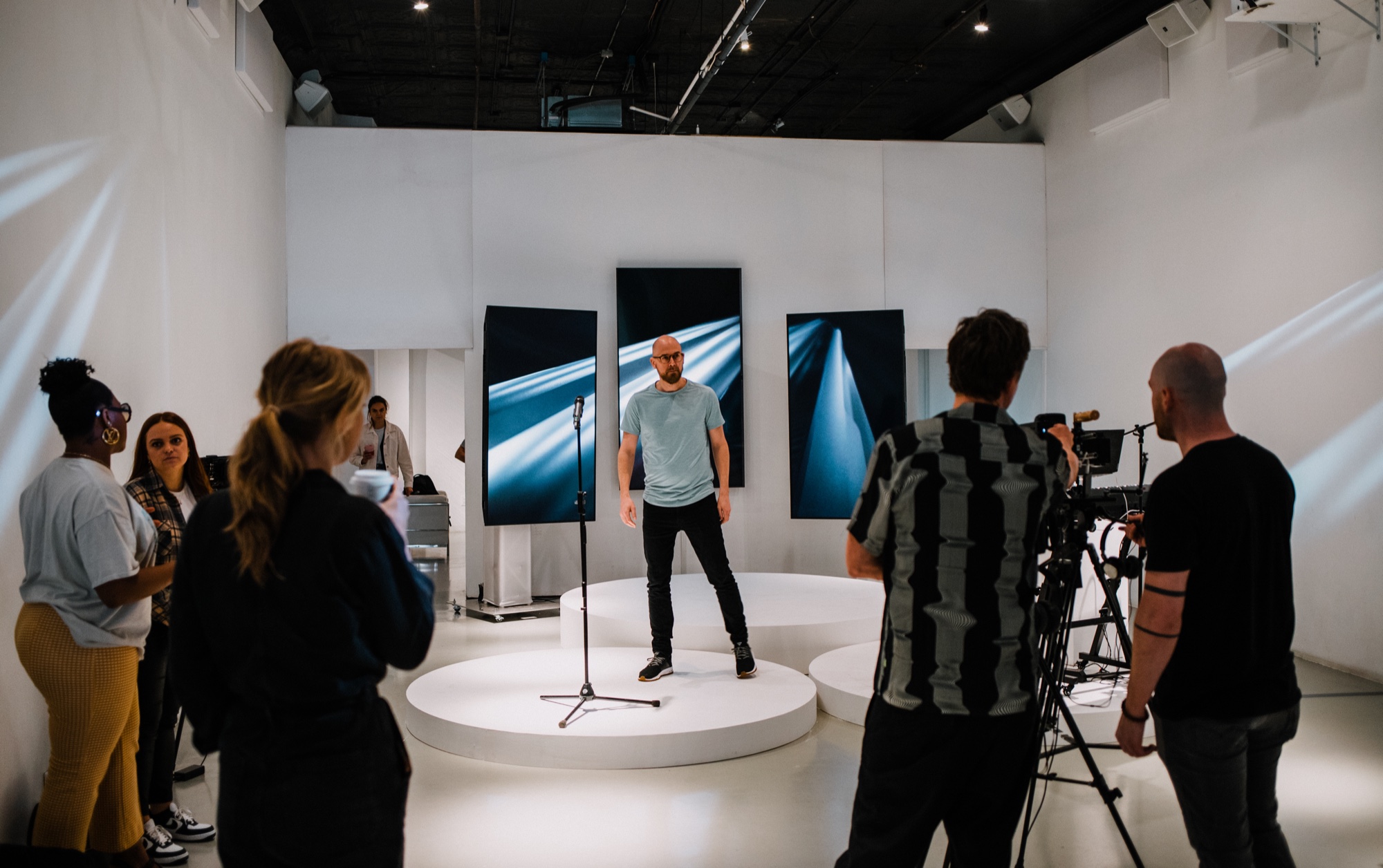 A man in a blue tee stands on a round white podium at a microphone while a crew films