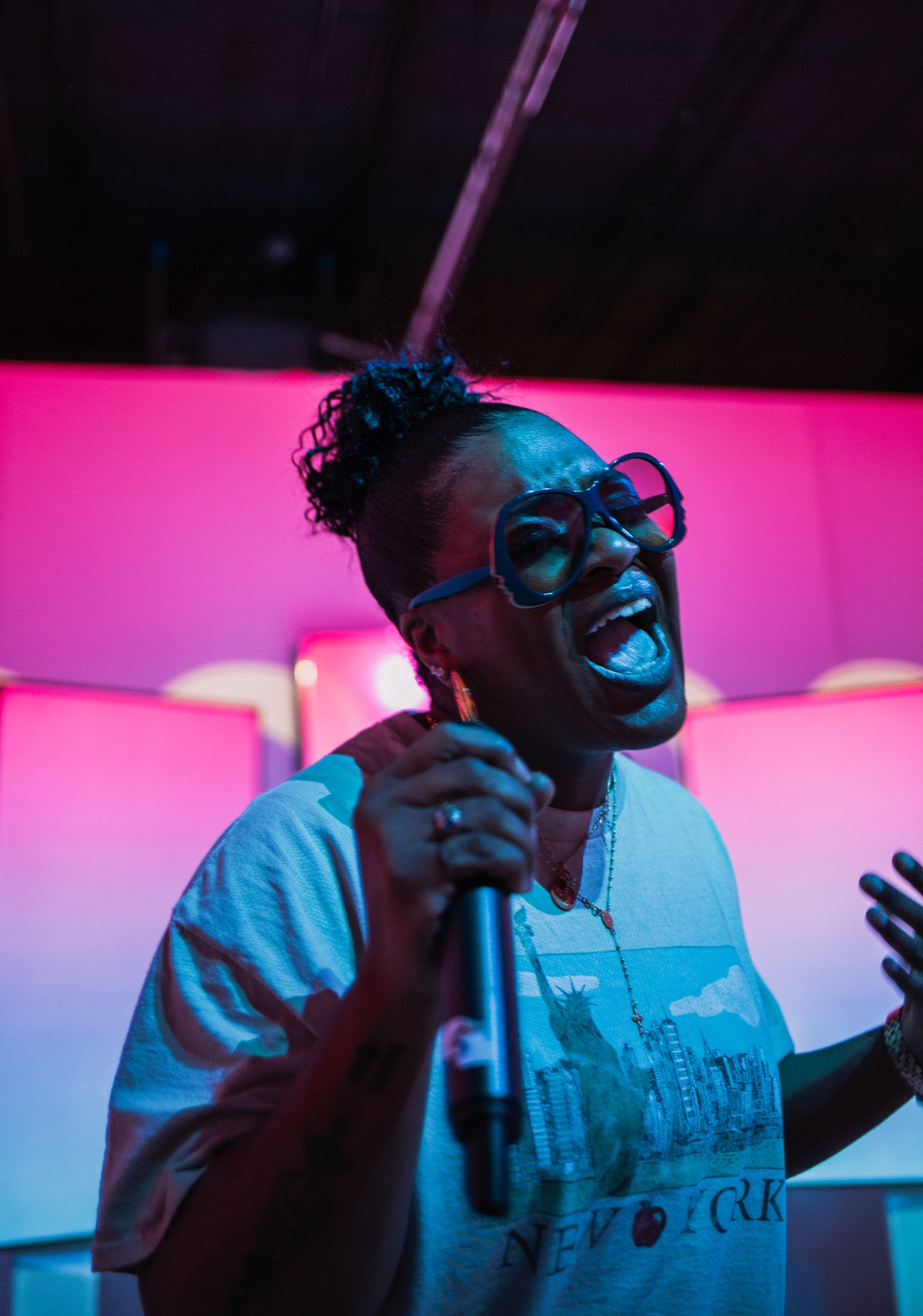 Woman in bold glasses singing into a microphone against a pink and blue lit backdrop