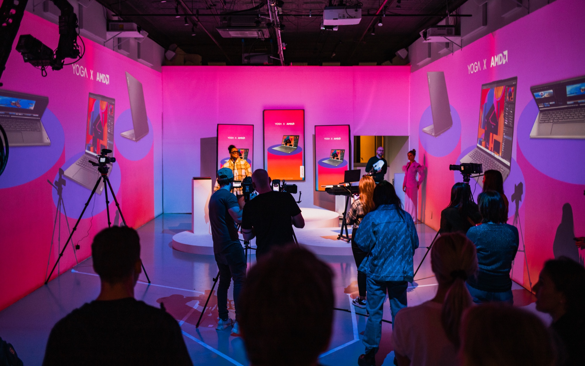 Camera crew films a person on a small stage in a pink and purple studio with Yoga x AMD posters