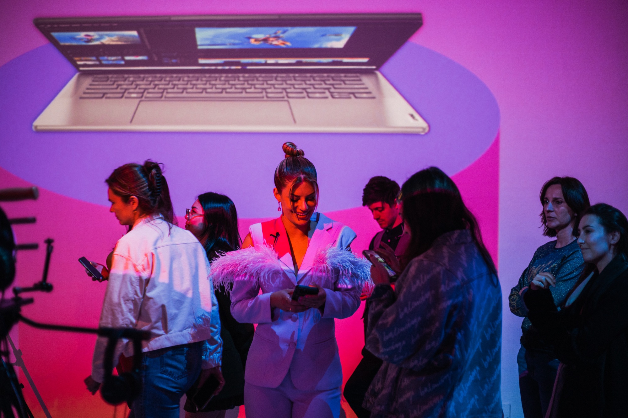 Guests check phones under a giant projected laptop, one woman in a white feathered suit at centre
