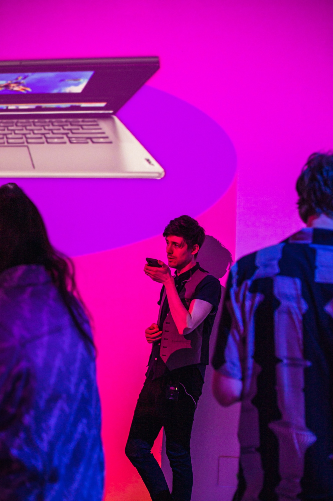 Man checking his phone against a magenta wall beneath a projected laptop image with guests in foreground