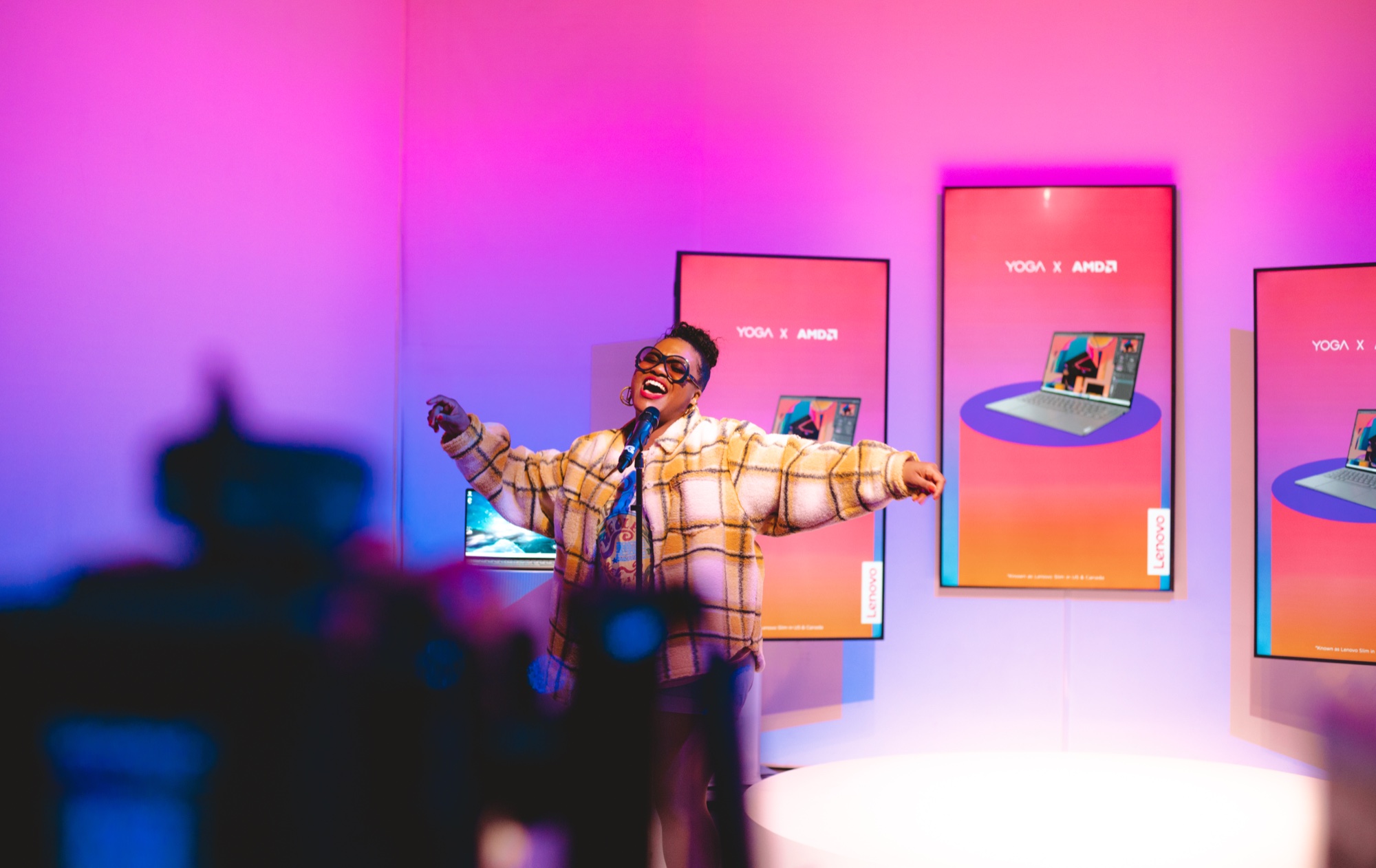 A performer in a plaid coat sings with arms spread on a pink and blue set beside Yoga AMD screens