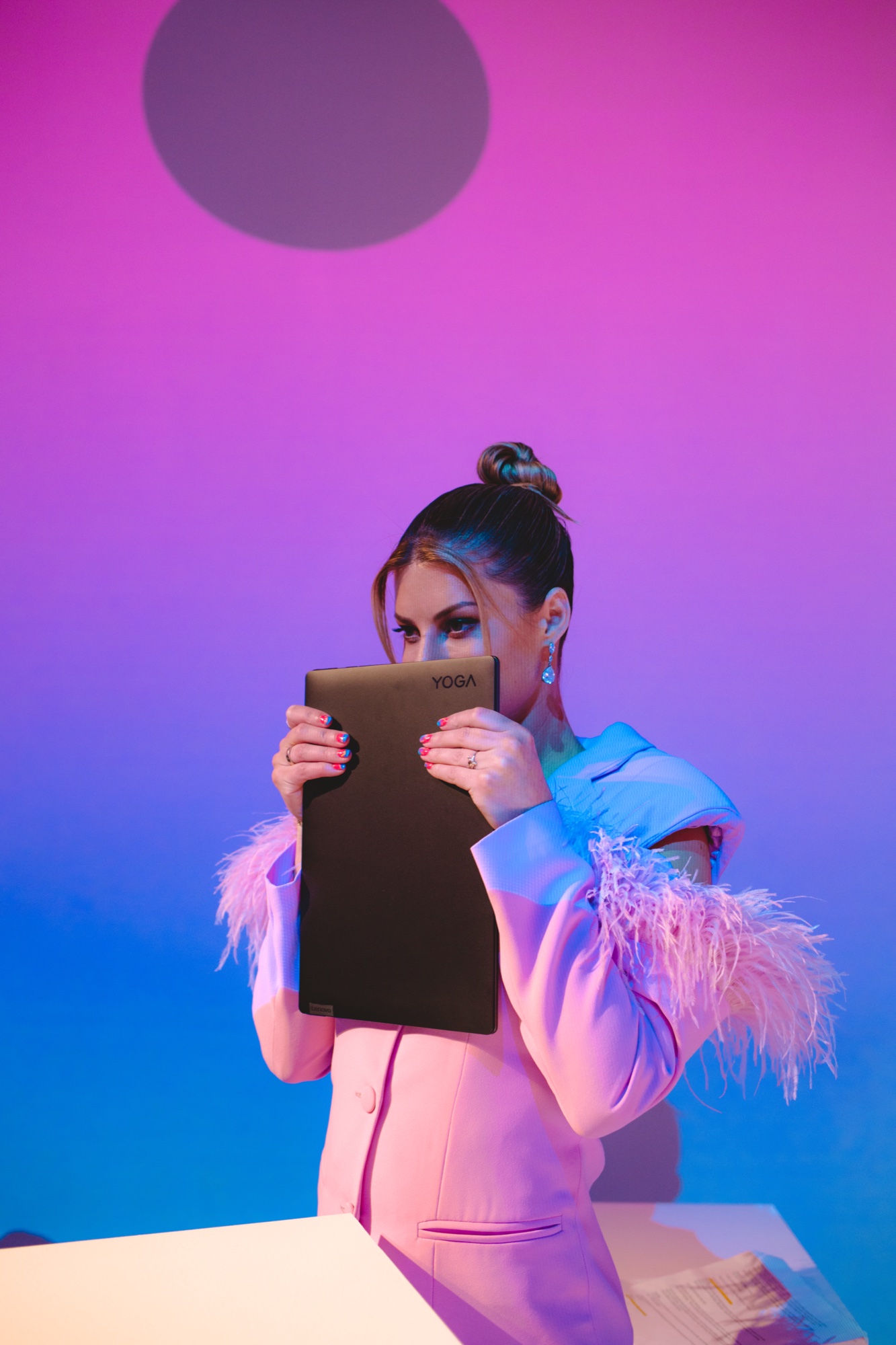 A woman in a pink feathered suit holds a Yoga laptop over her face against a purple and blue wall