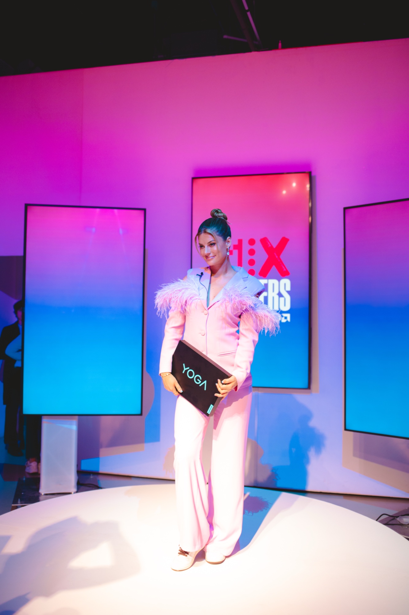 A presenter in a pale pink feathered suit holds a Yoga laptop on a round white stage before screens