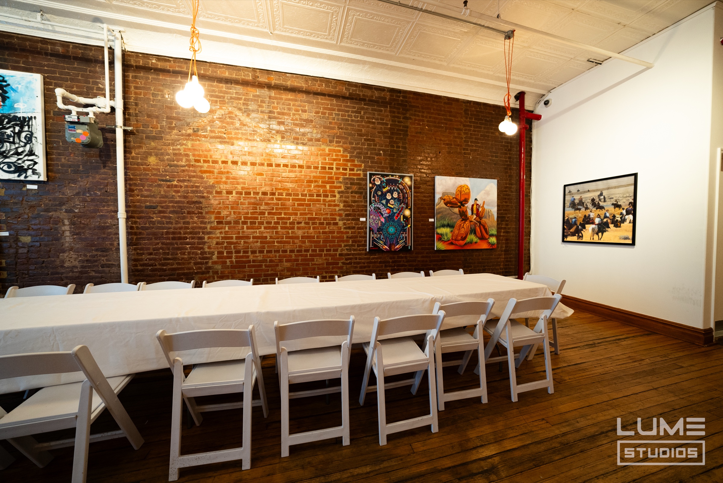 Long white draped dinner table with white folding chairs beside an exposed brick wall hung with paintings