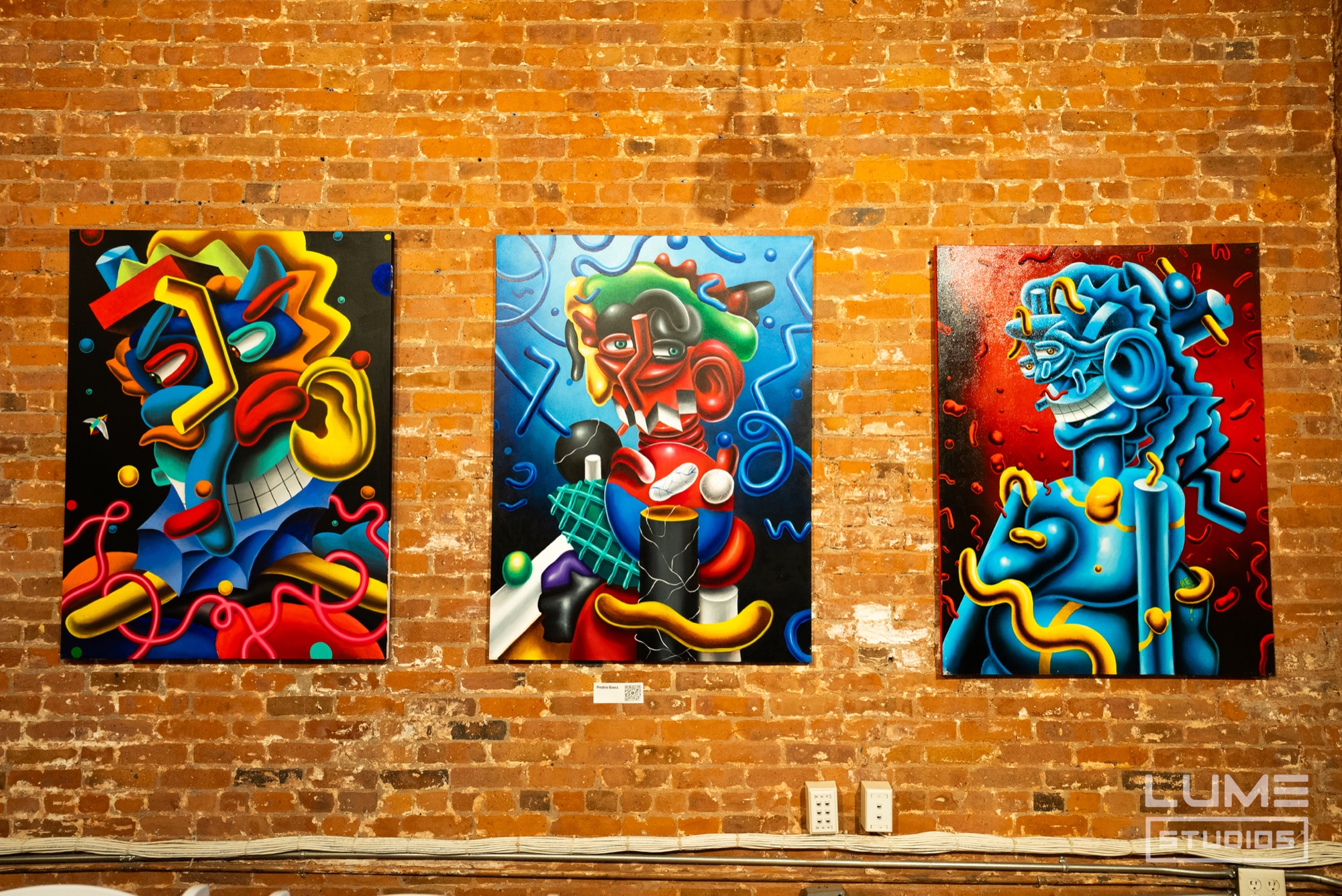 Three colourful cartoon style paintings in blue, red and yellow hung on an exposed brick wall