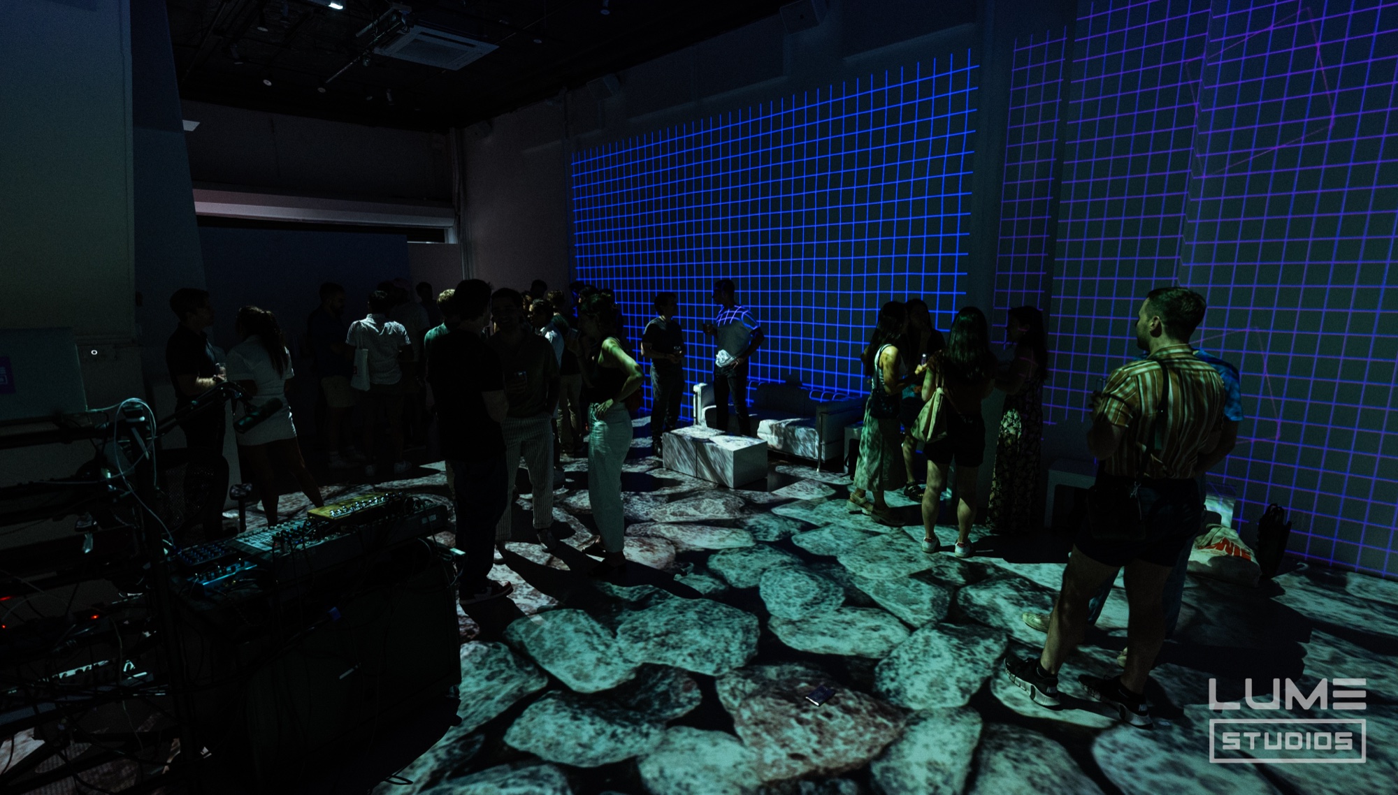 Guests stand on a floor projected with grey stones beneath a blue grid wall in a dark room