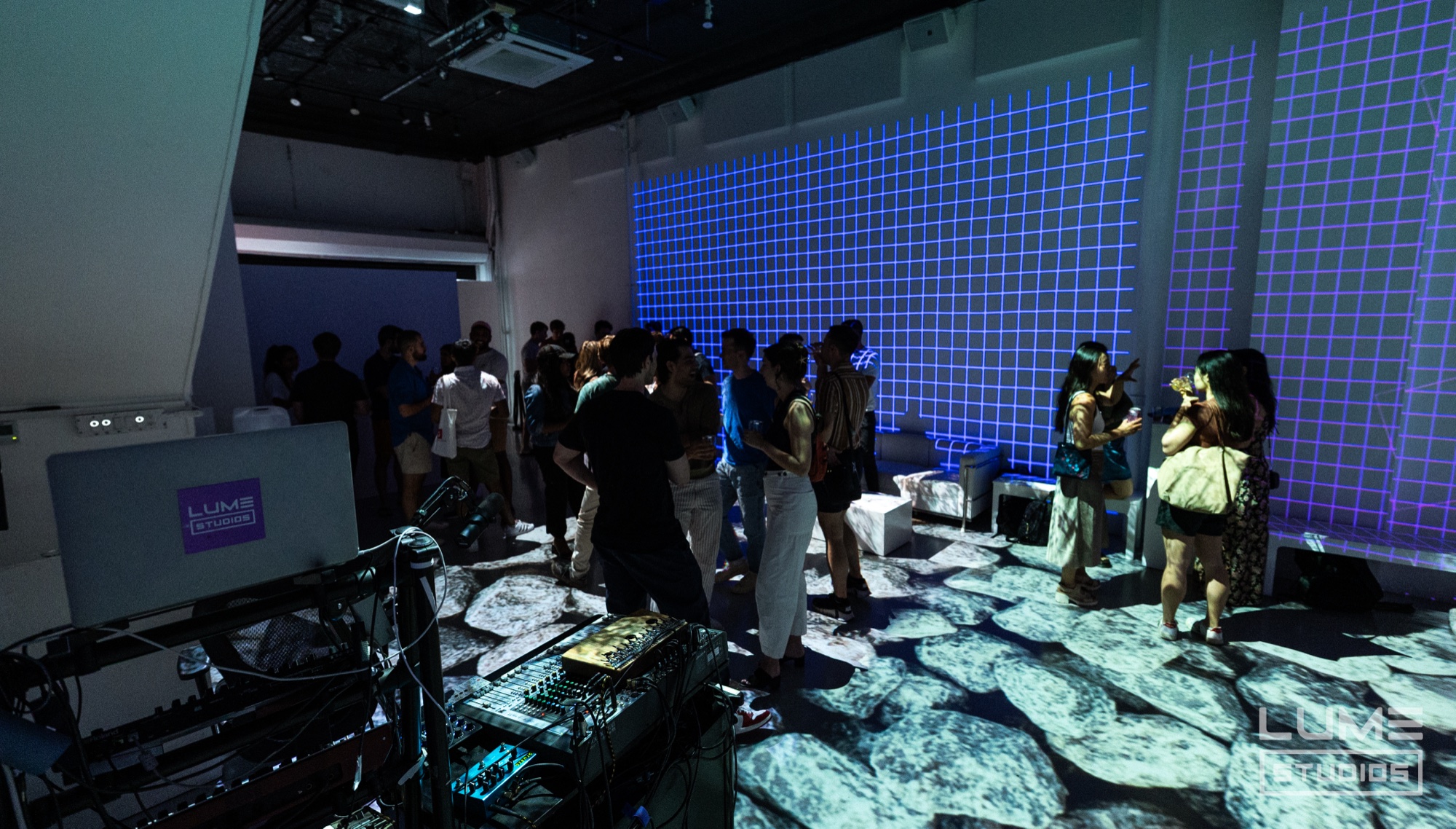 Guests stand on a floor projected with cracked ice beneath a blue grid wall, DJ gear in the foreground