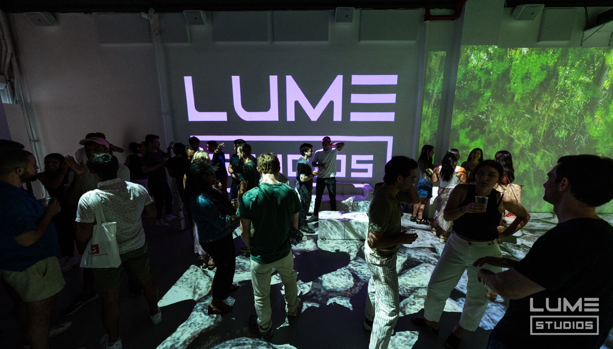 Crowd in a dark room facing a purple Lume Studios logo projection beside green forest imagery
