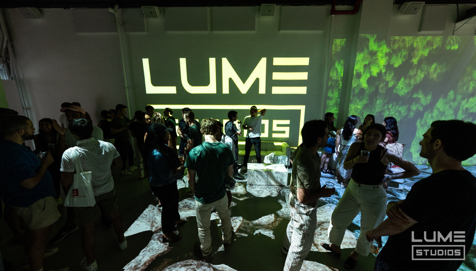 Crowd gathers under a large yellow Lume logo projected on the wall, green foliage projected at right