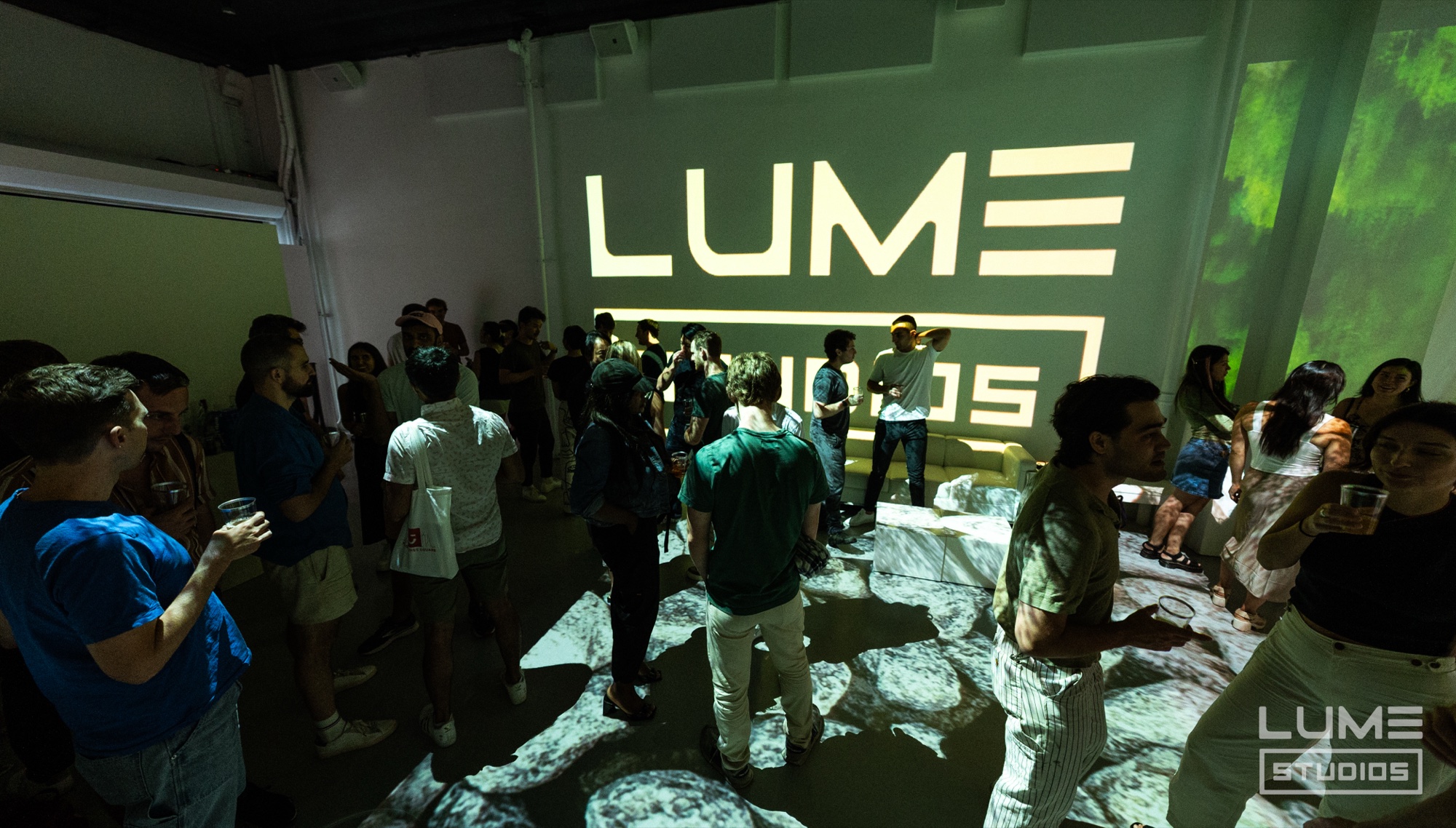 Crowd stands in a green lit room with a large Lume Studios logo projected on the wall