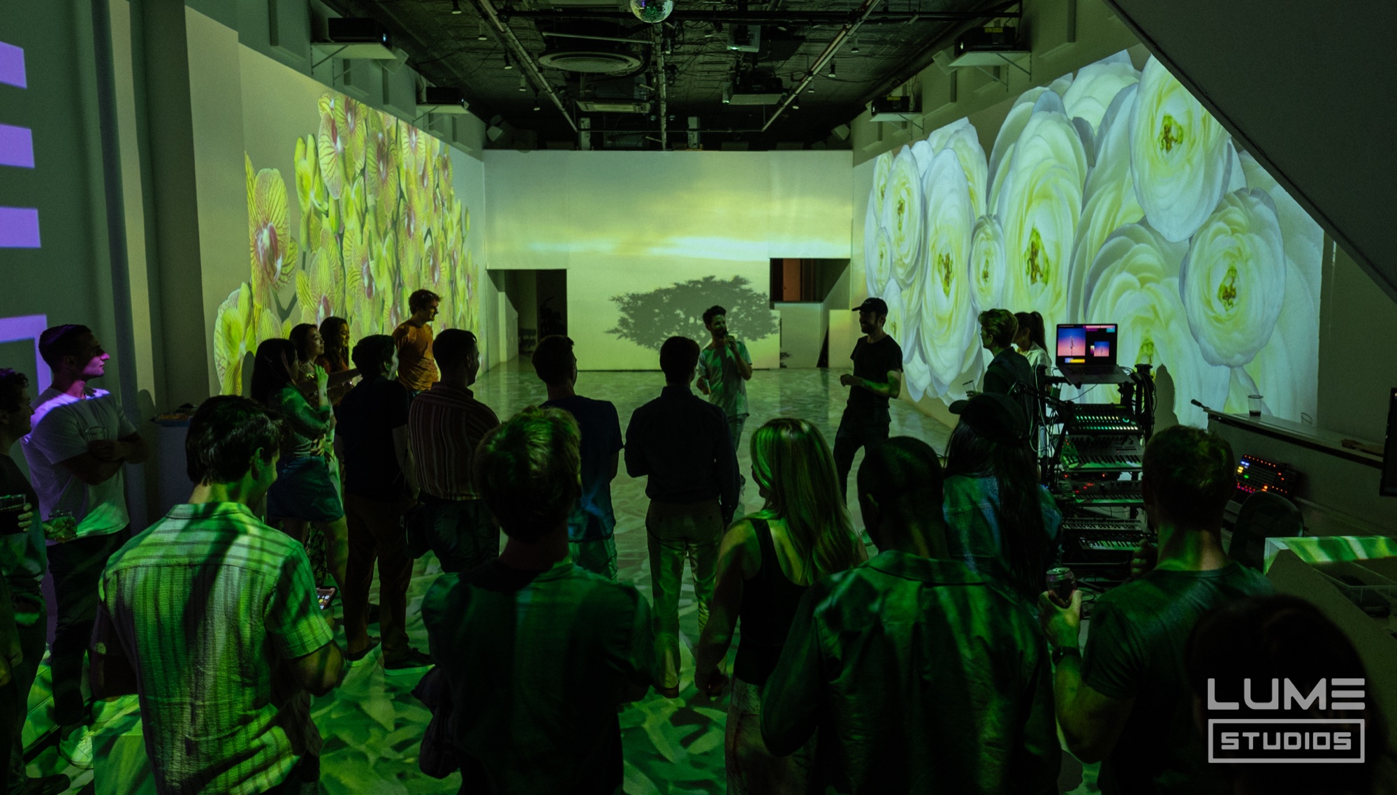 Crowd in green light watches a speaker before walls projected with white roses