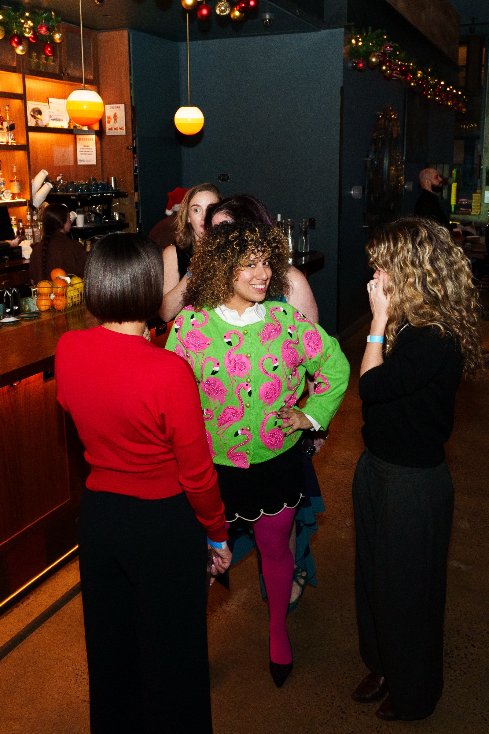A woman in a green cardigan printed with pink flamingos chats with guests beside a bar hung with holiday garland