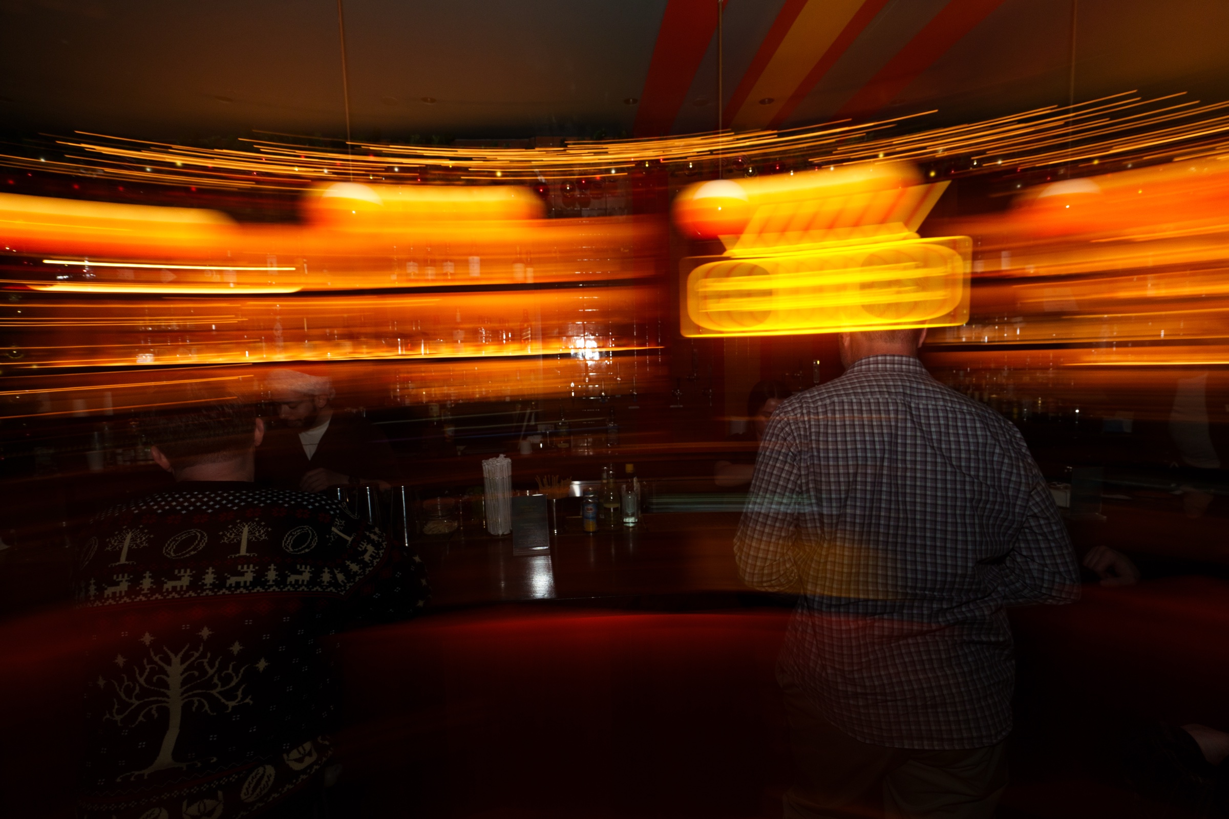 Long exposure of a bar with streaks of orange light behind a man in a checked shirt at a table
