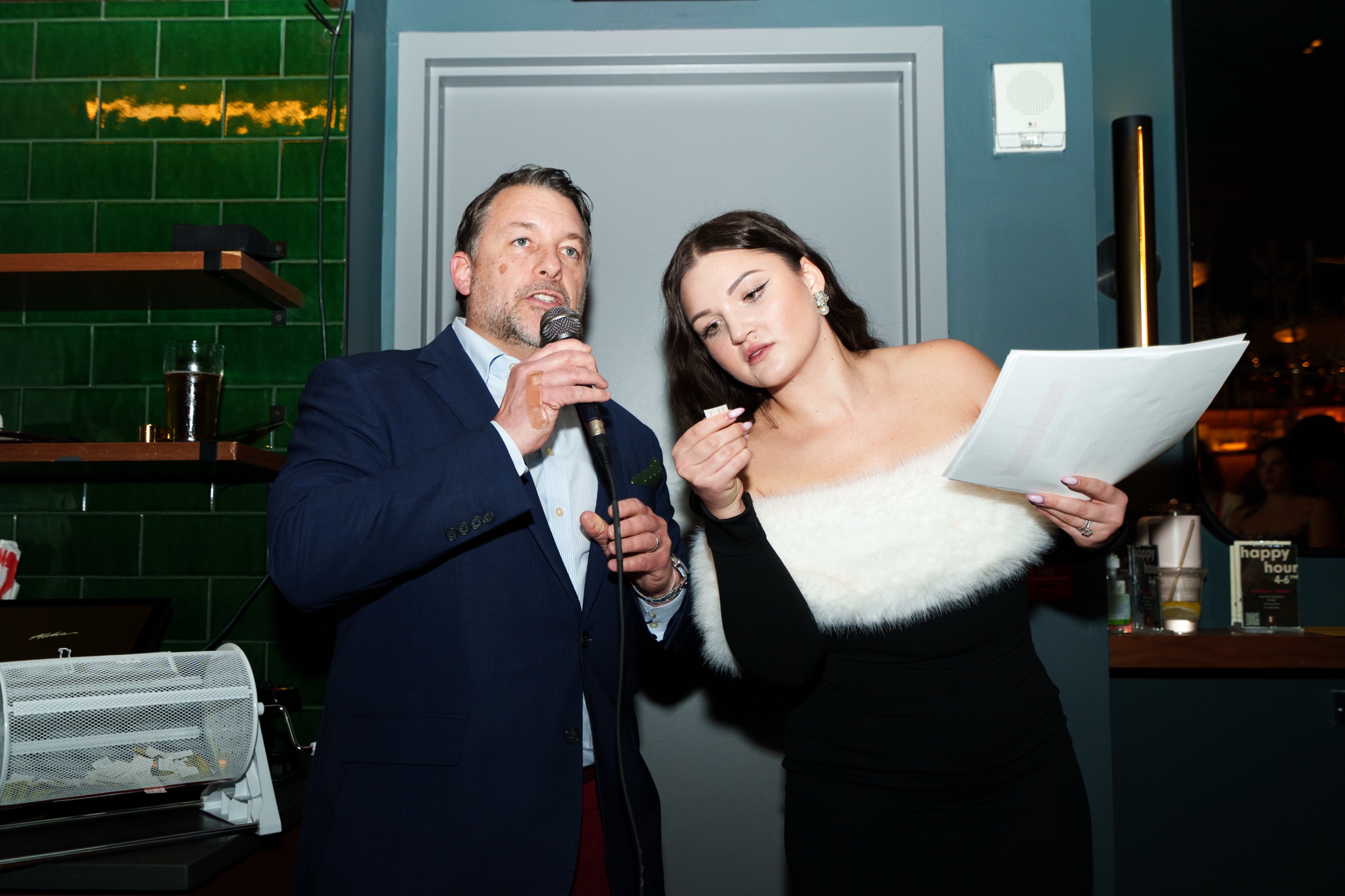 A man in a navy blazer speaks into a microphone while a woman in a white fur wrap reads notes