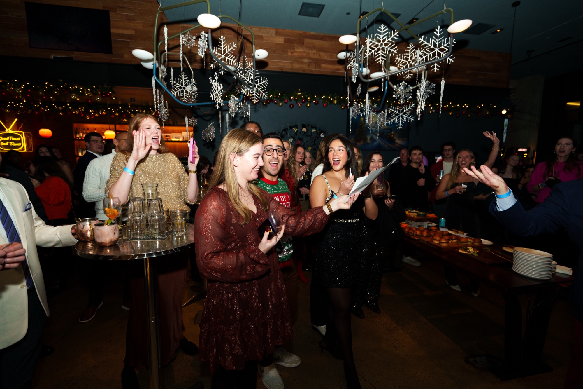 Guests singing and holding drinks beneath hanging snowflake decorations at a festive bar