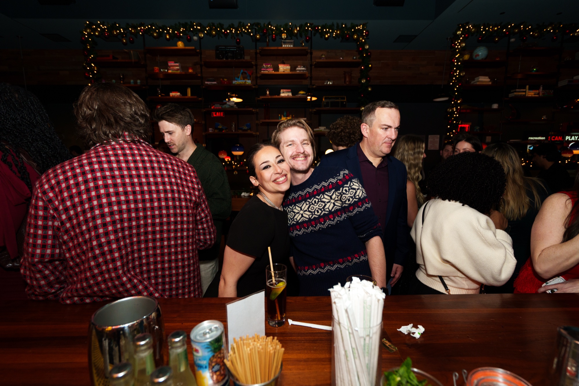 Three friends smile at a bar counter beneath string lights and garland, one in a patterned holiday sweater