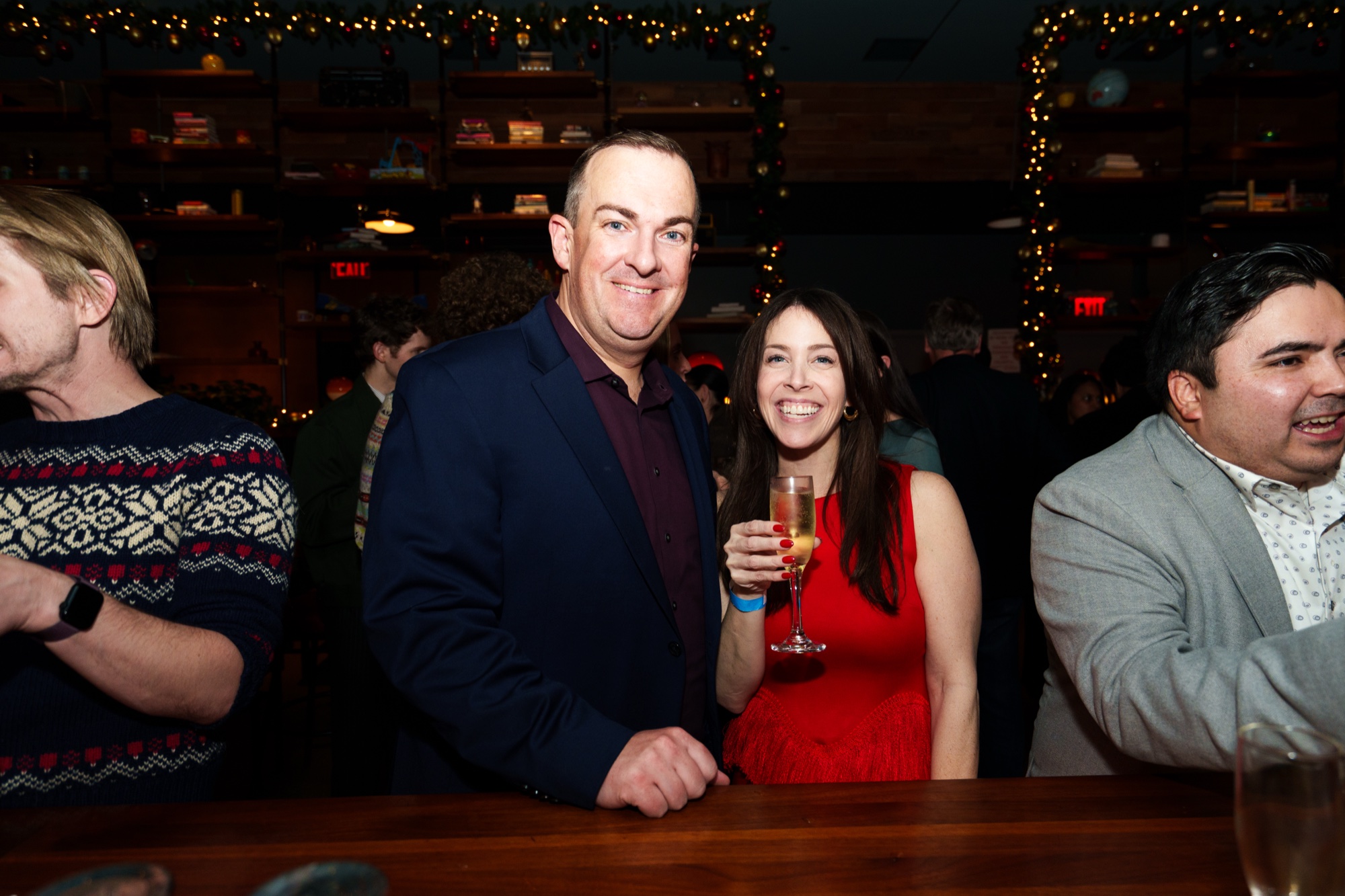 A man in a navy blazer and a woman in red holding champagne smile at a string lit bar