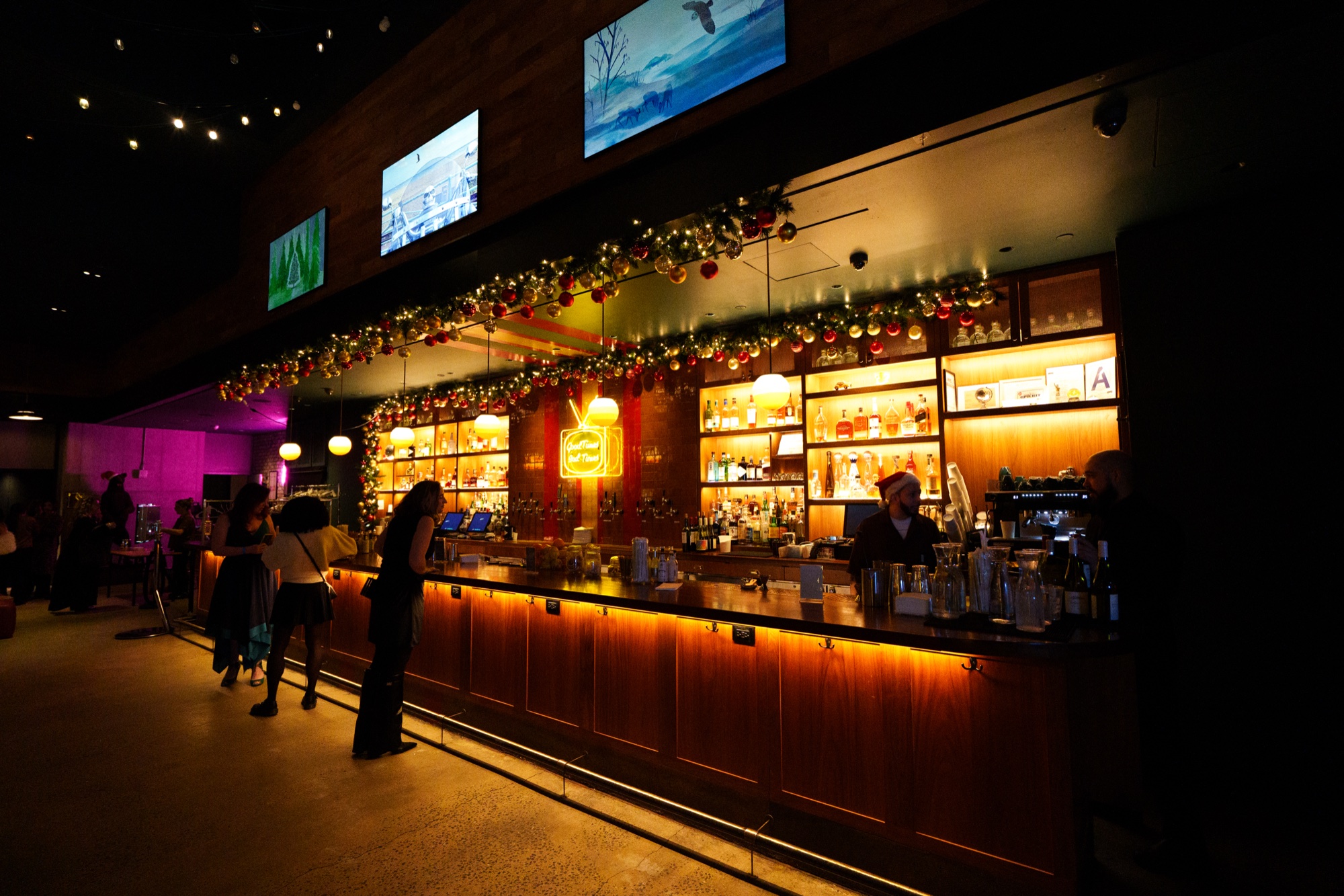 Long wooden bar lined with garlands and warm backlit shelves of bottles below wall mounted screens