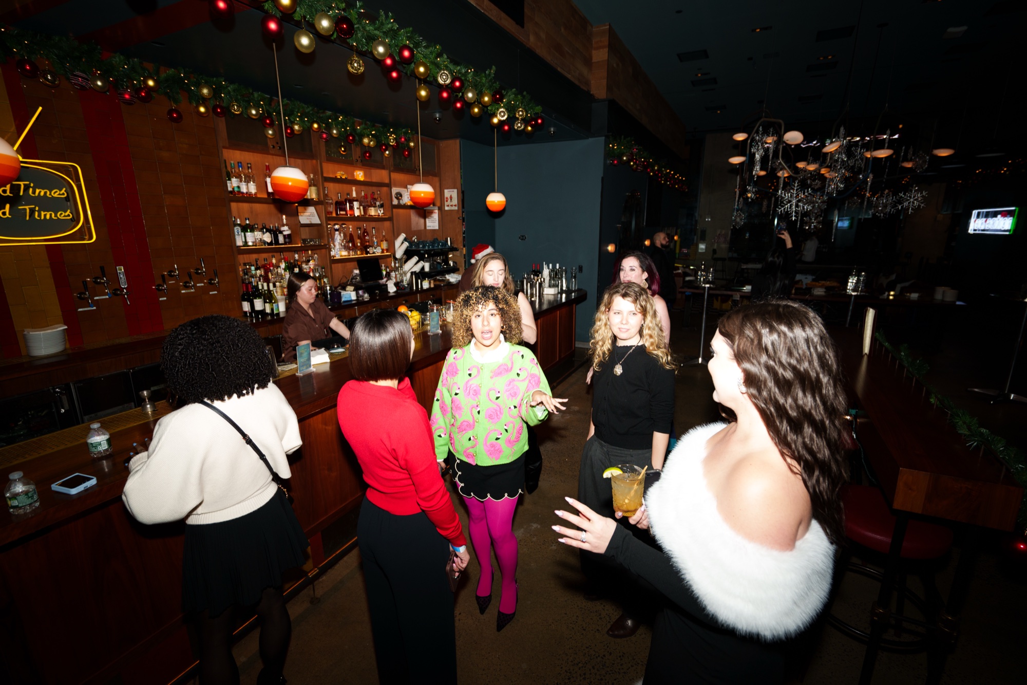 Guests in party outfits chat in front of a wooden bar trimmed with garland and orange lanterns