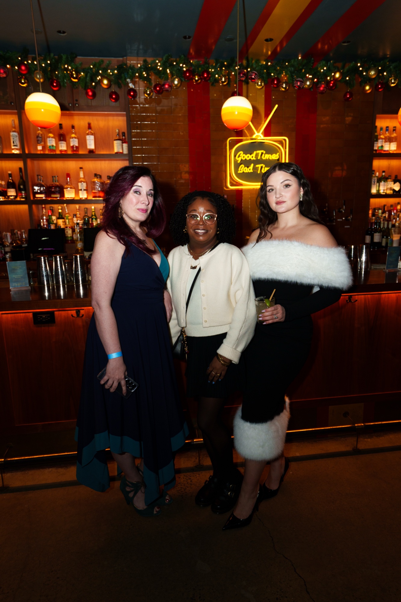 Three women pose at a wood panelled bar with holiday garland and a yellow neon sign reading Good Times