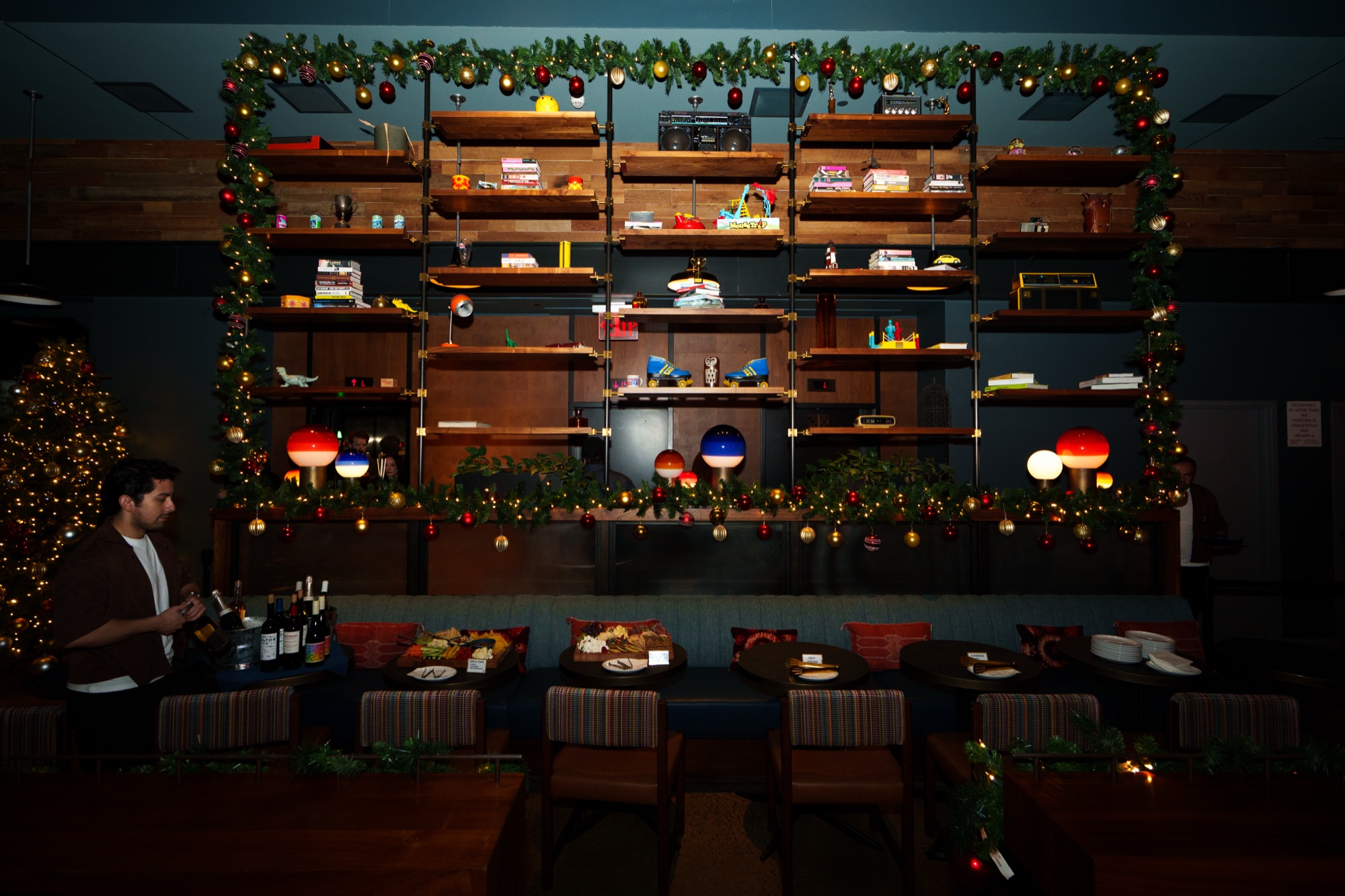 A wall of wooden shelves wrapped in holiday garlands and lanterns above dining booths, a bartender at left