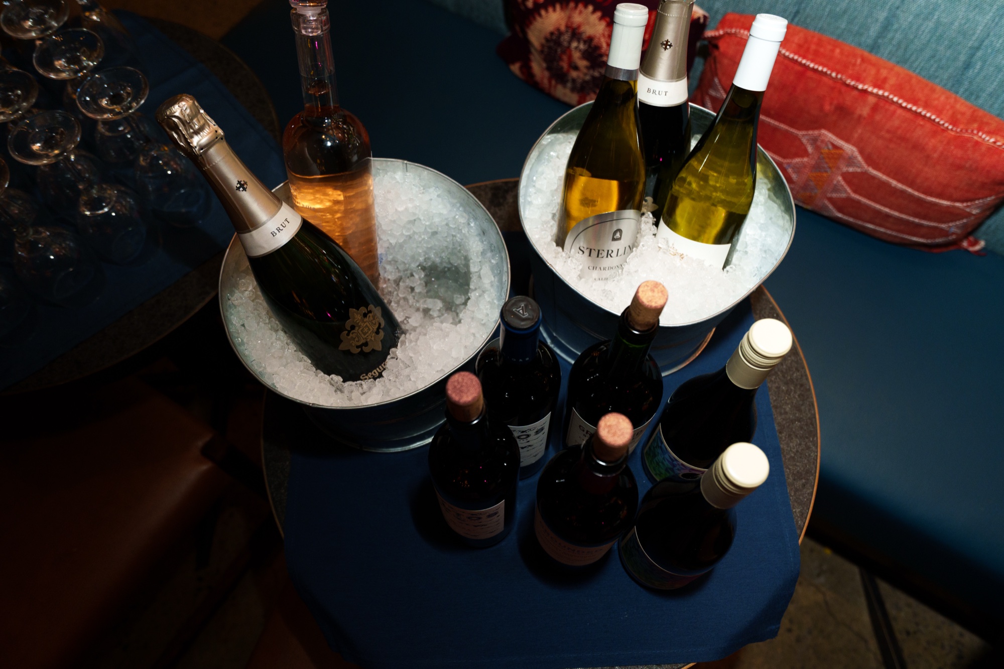 Bottles of champagne and wine sit in ice buckets on a blue table beside a red patterned cushion
