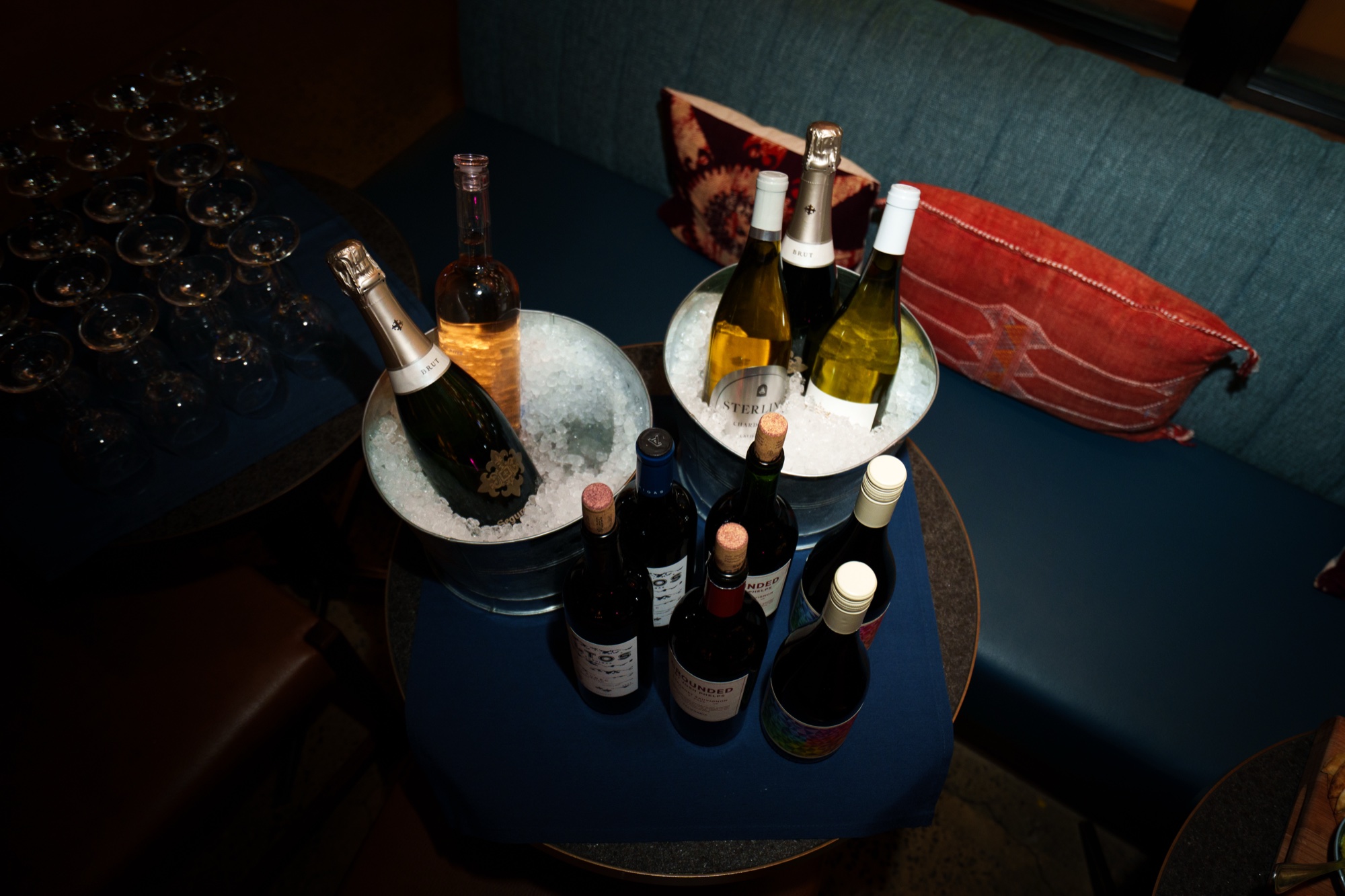 Champagne and wine bottles in ice buckets on a small blue table beside a booth with a red cushion