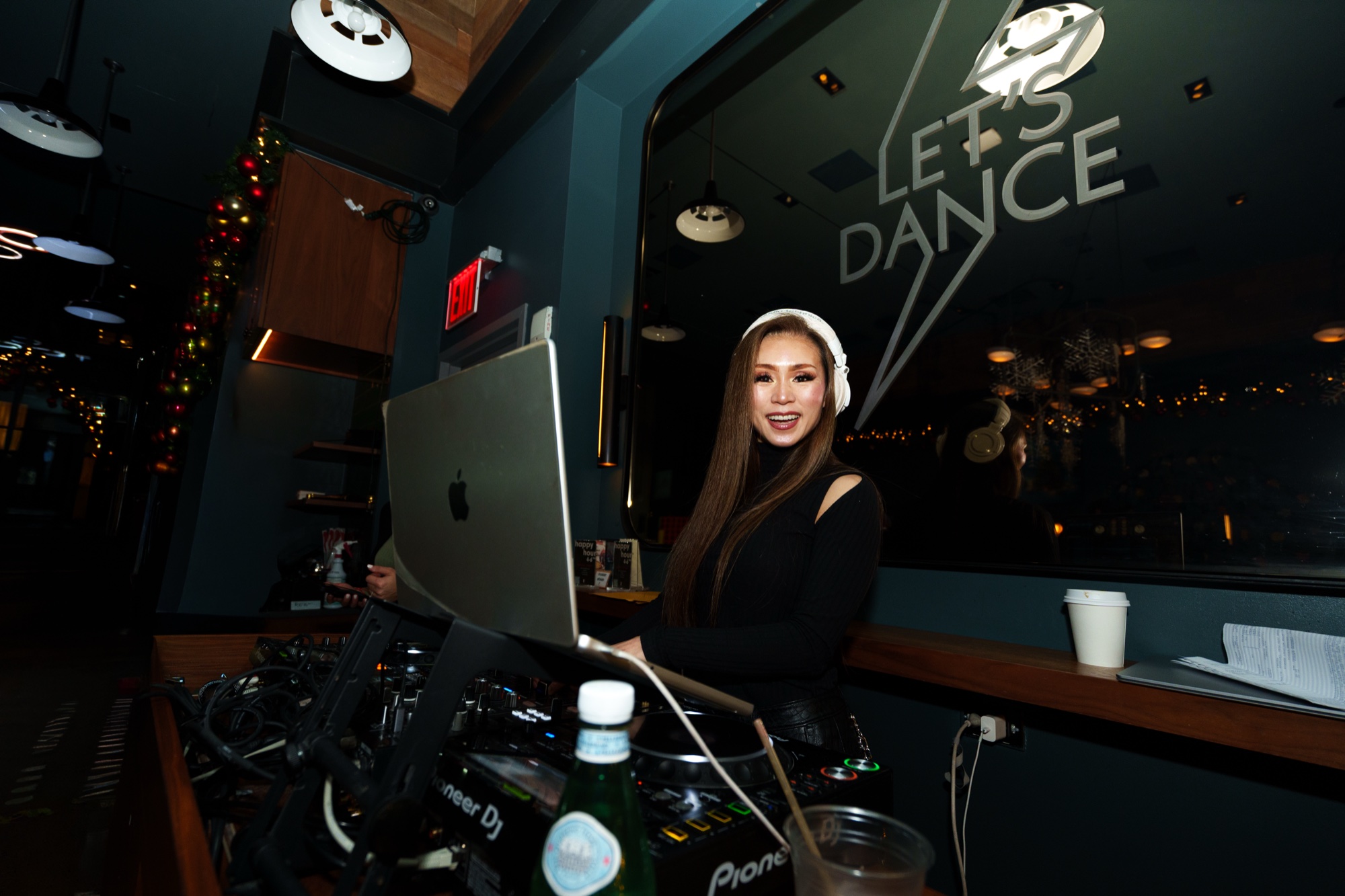 Smiling DJ in headphones behind a laptop and decks before a window reading let’s dance