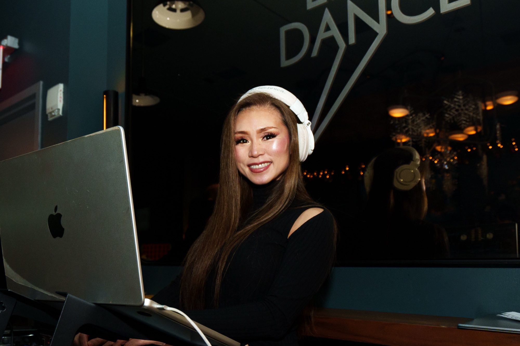 A DJ in white headphones smiles behind a laptop in front of a window sign reading Dance
