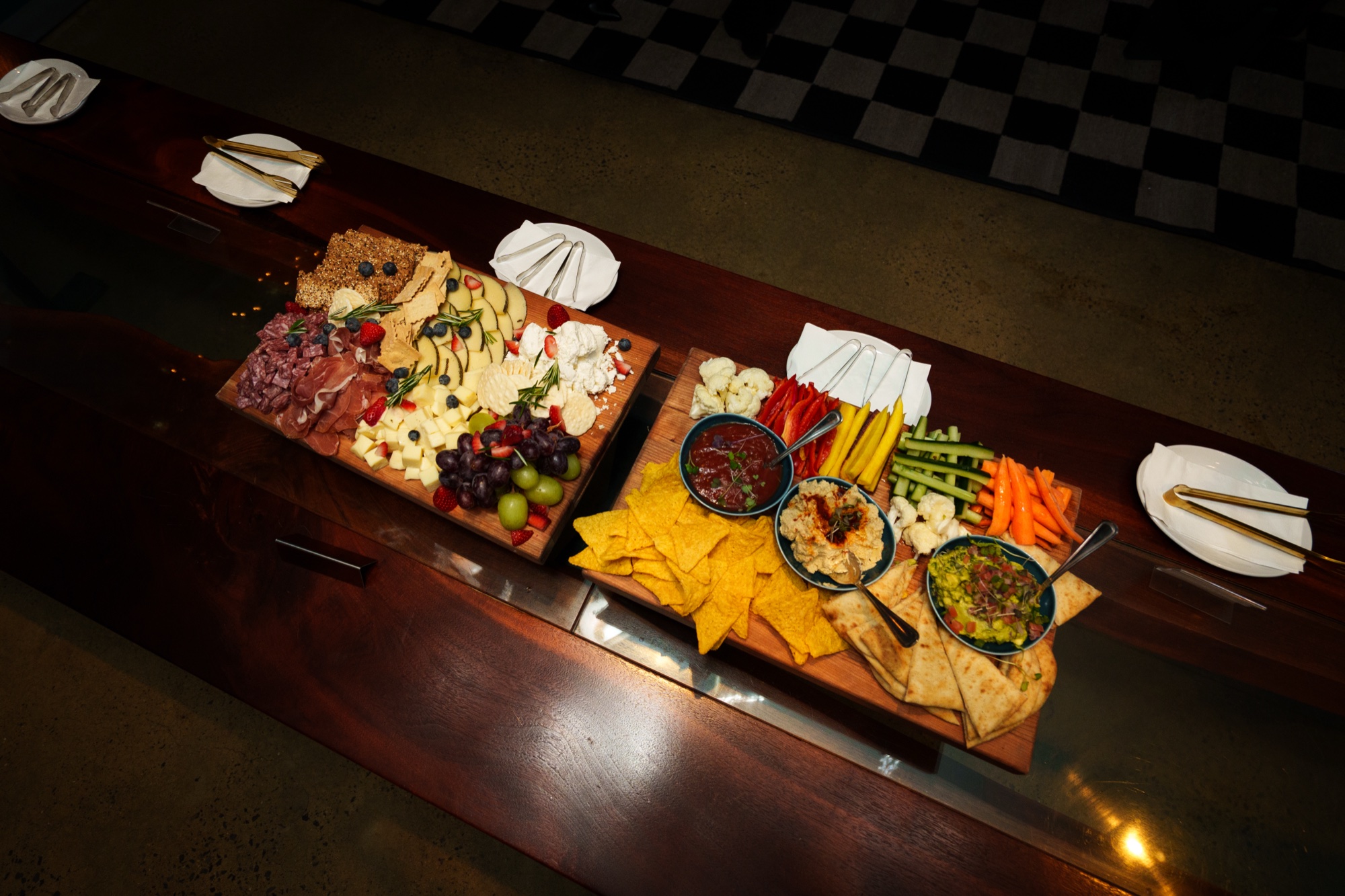 Two wooden grazing boards of cheese, cured meats, chips and vegetable dips on a long dark table