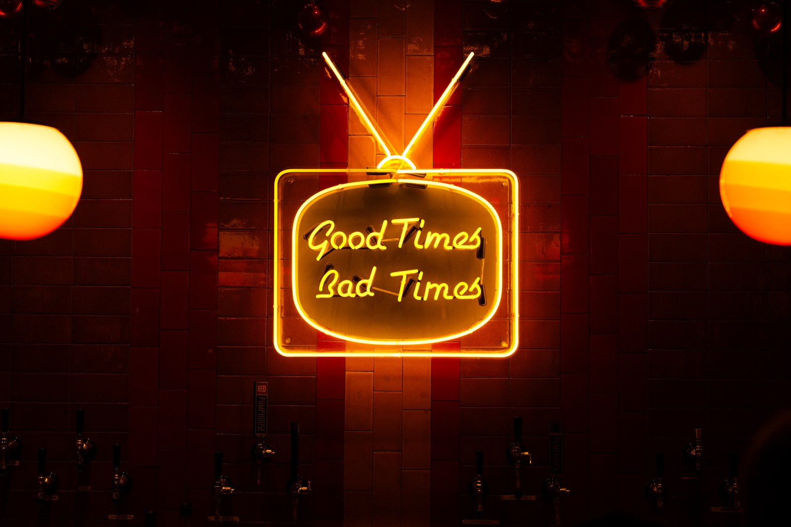 A neon sign shaped like a television, reading Good Times Bad Times in orange script, glowing on the tiled wall above the bar