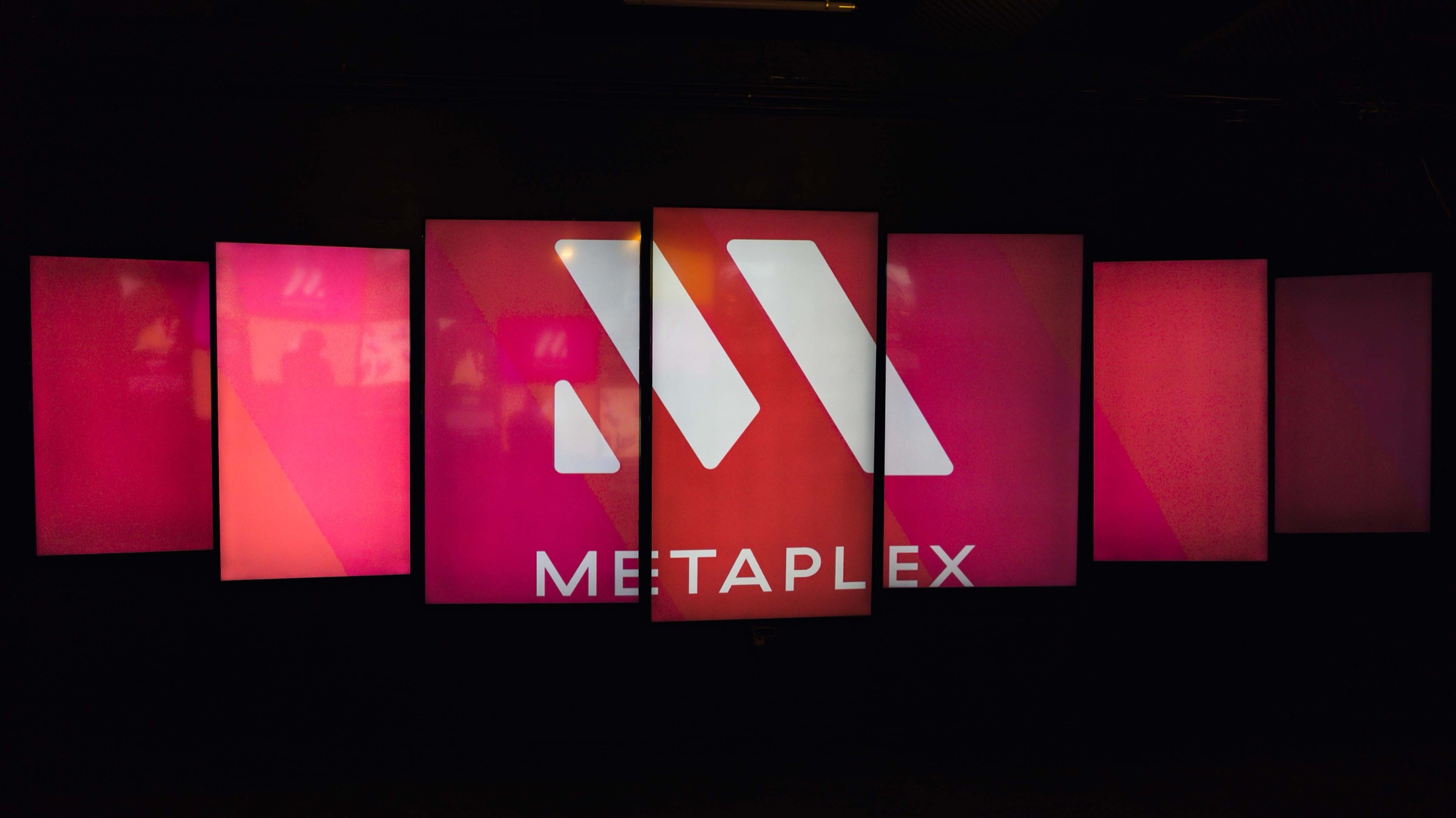 Metaplex logo glows white across a row of pink and red vertical screens in a dark room