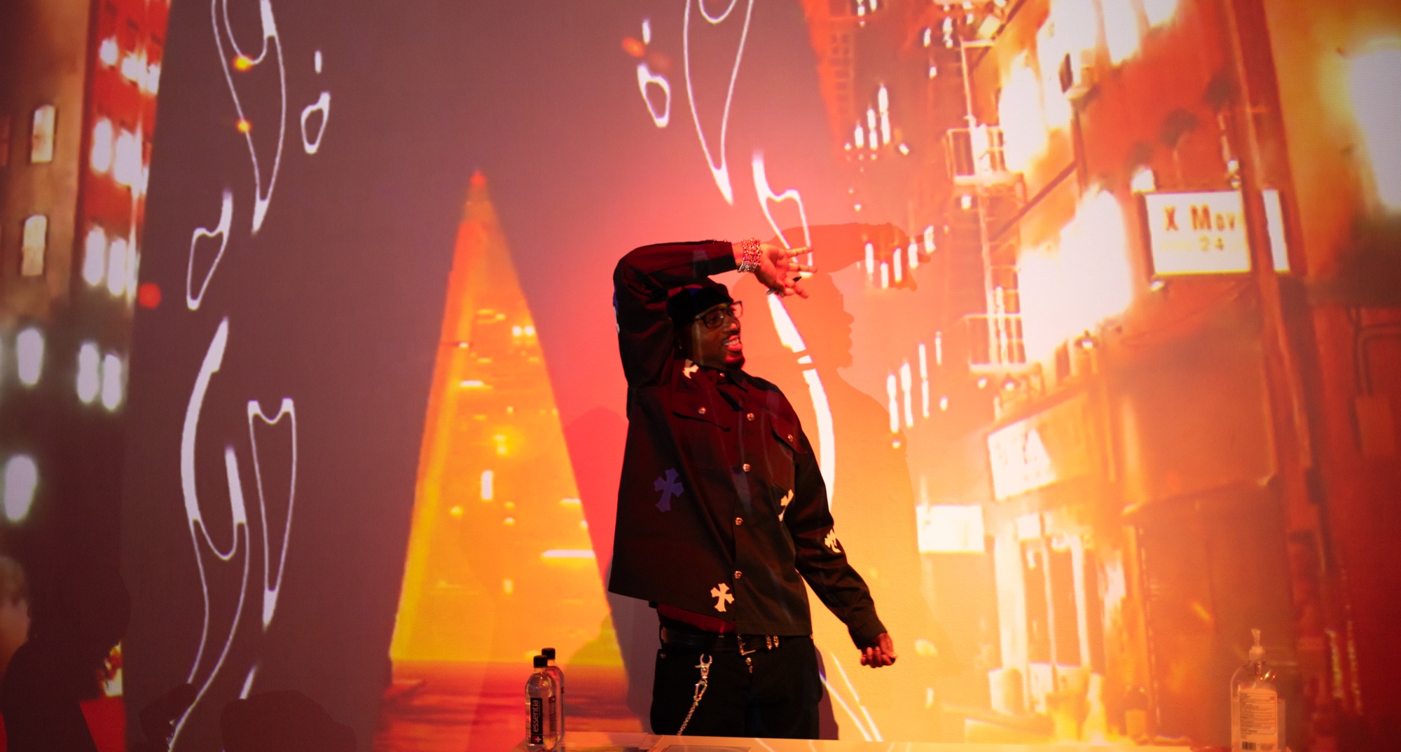 A man in a dark jacket raises an arm before a wall projected with orange city streets and line drawings