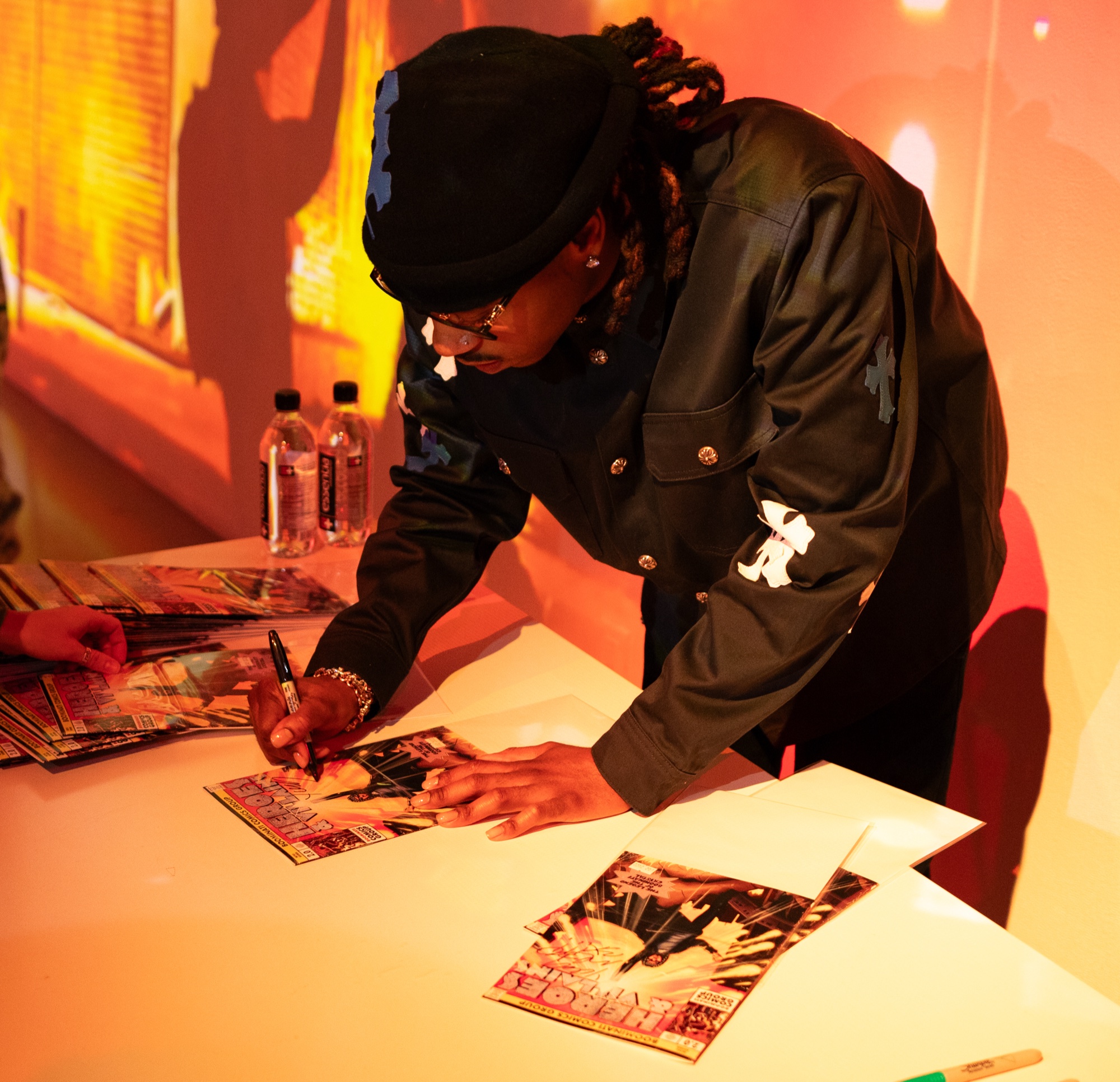 A man in a black jacket and beanie signs comic book covers at a white table in warm orange light