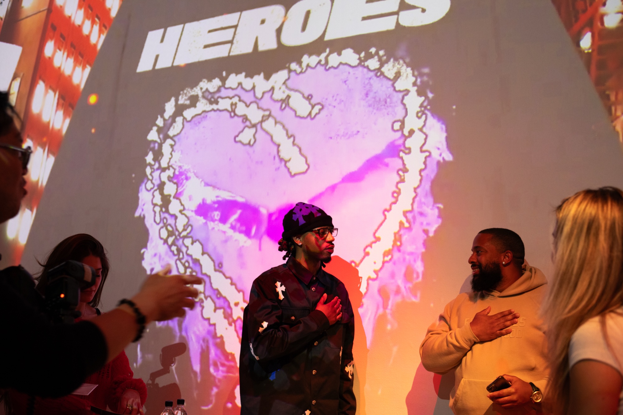 A man in a black beanie and cross patched jacket stands before a projected flaming heart and the word Heroes
