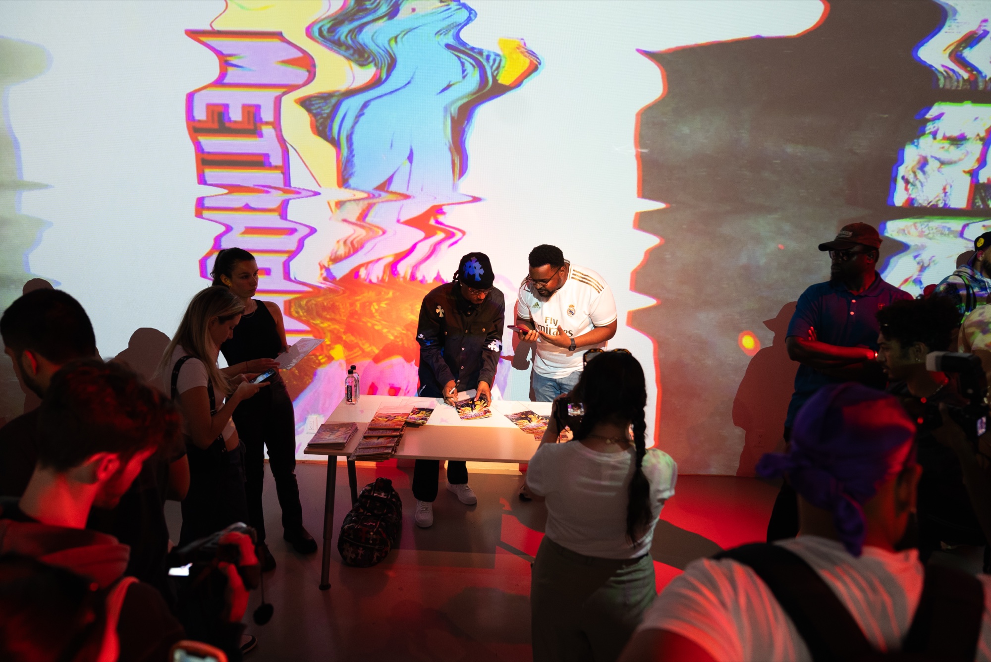 Guests watch two people signing at a table beneath a wall projection of glitched Metro lettering