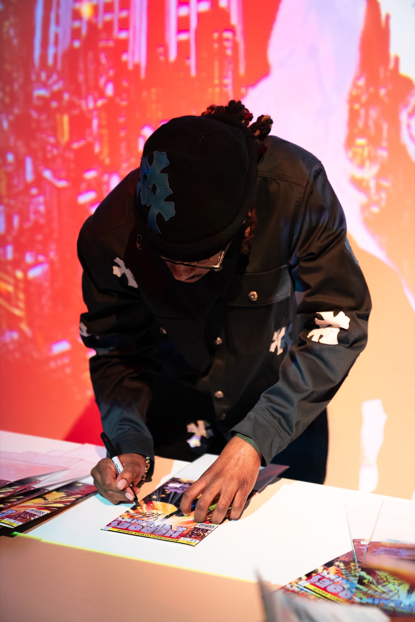 A man in a black beanie and glasses signs a comic style print before a red projected cityscape