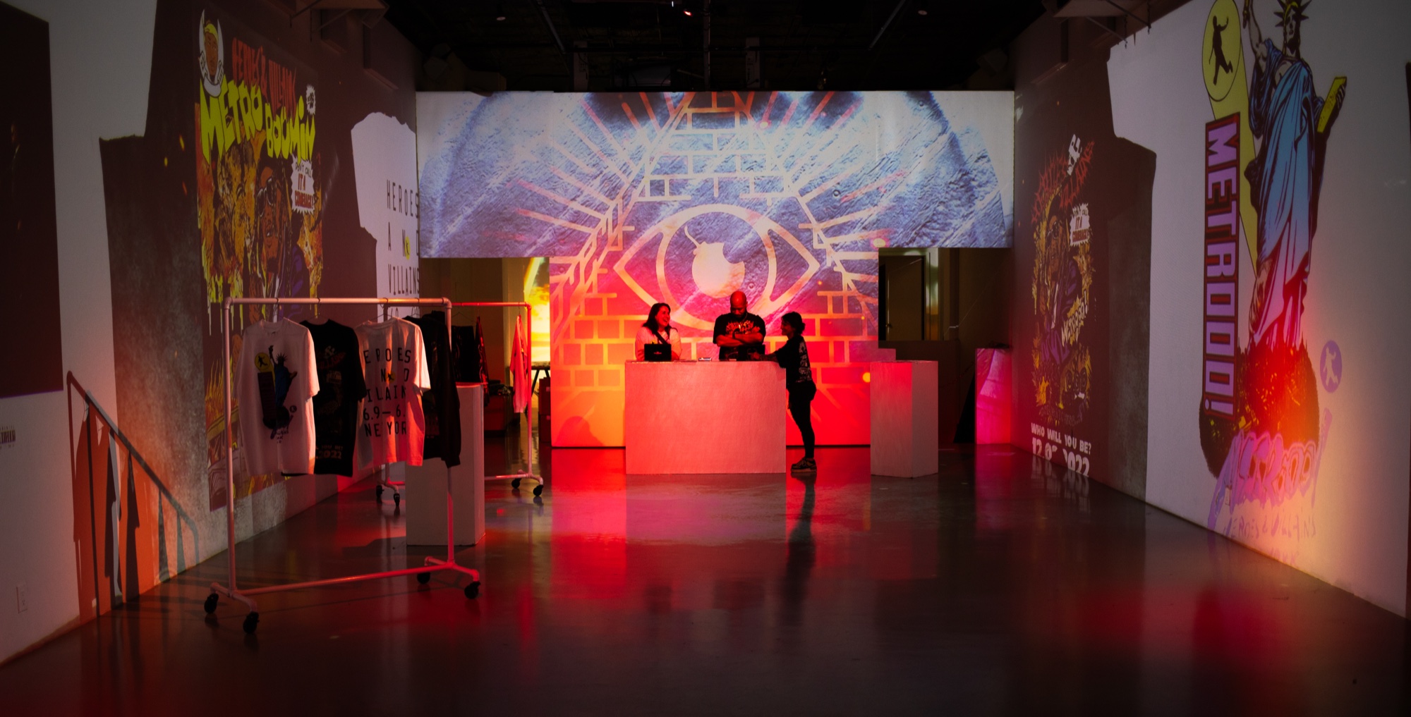 Two people at a white counter beneath a projected pyramid eye mural with a rack of shirts and red light
